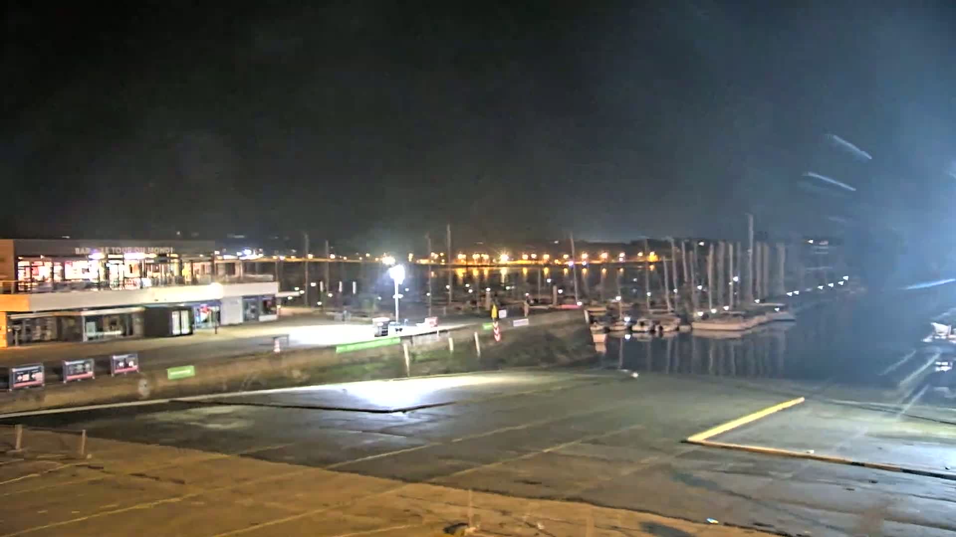 A nighttime view of a harbor with docked sailboats, a building with lights on, and an empty paved area in front.