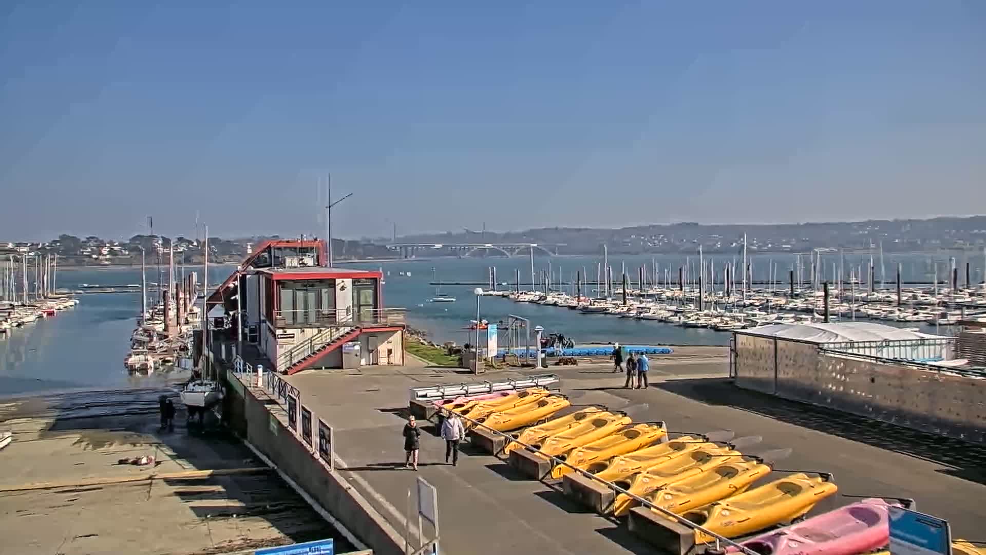 A marina with numerous sailboats and kayaks, a red building, and people walking on a sunny day.