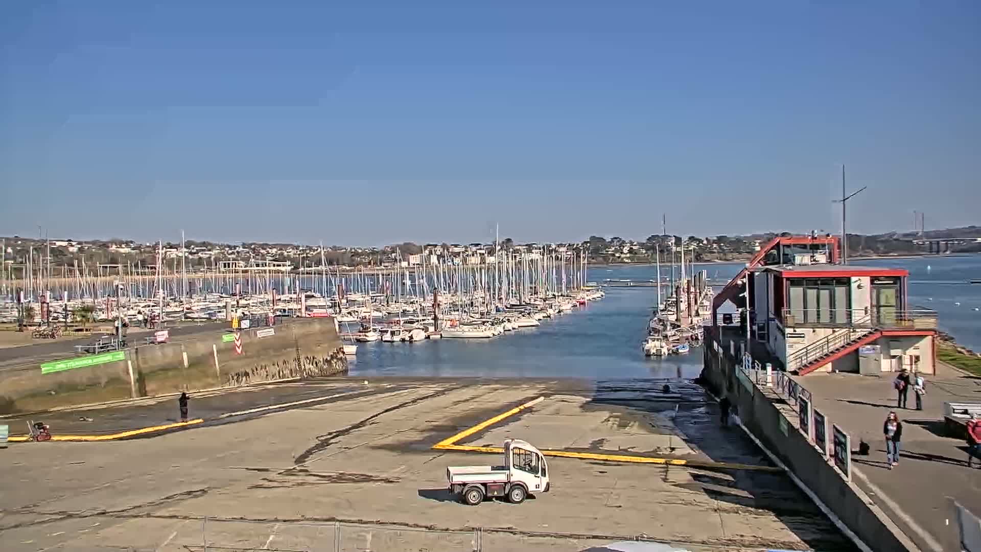 A marina filled with numerous sailboats under a clear blue sky is viewed from a high vantage point, with a small utility truck parked in the foreground near a building.