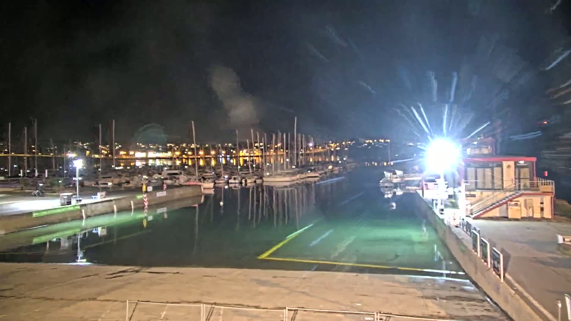 A nighttime view of a marina with numerous boats moored, a brightly lit area near a small building, and calm, greenish water reflecting the lights of the city in the background.