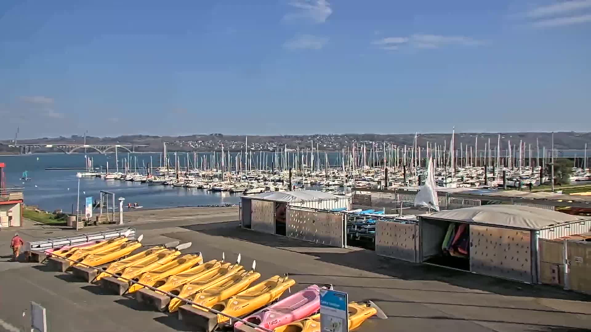A marina filled with sailboats under a mostly sunny sky, with rows of kayaks lined up on the shore.