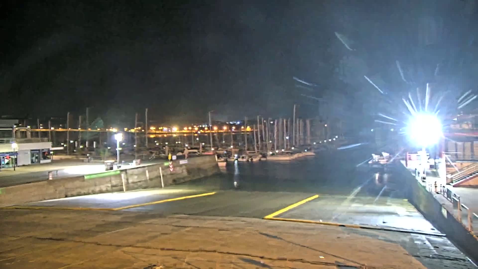 At night, a brightly lit boat ramp leads to a harbor filled with docked sailboats under a clear sky.