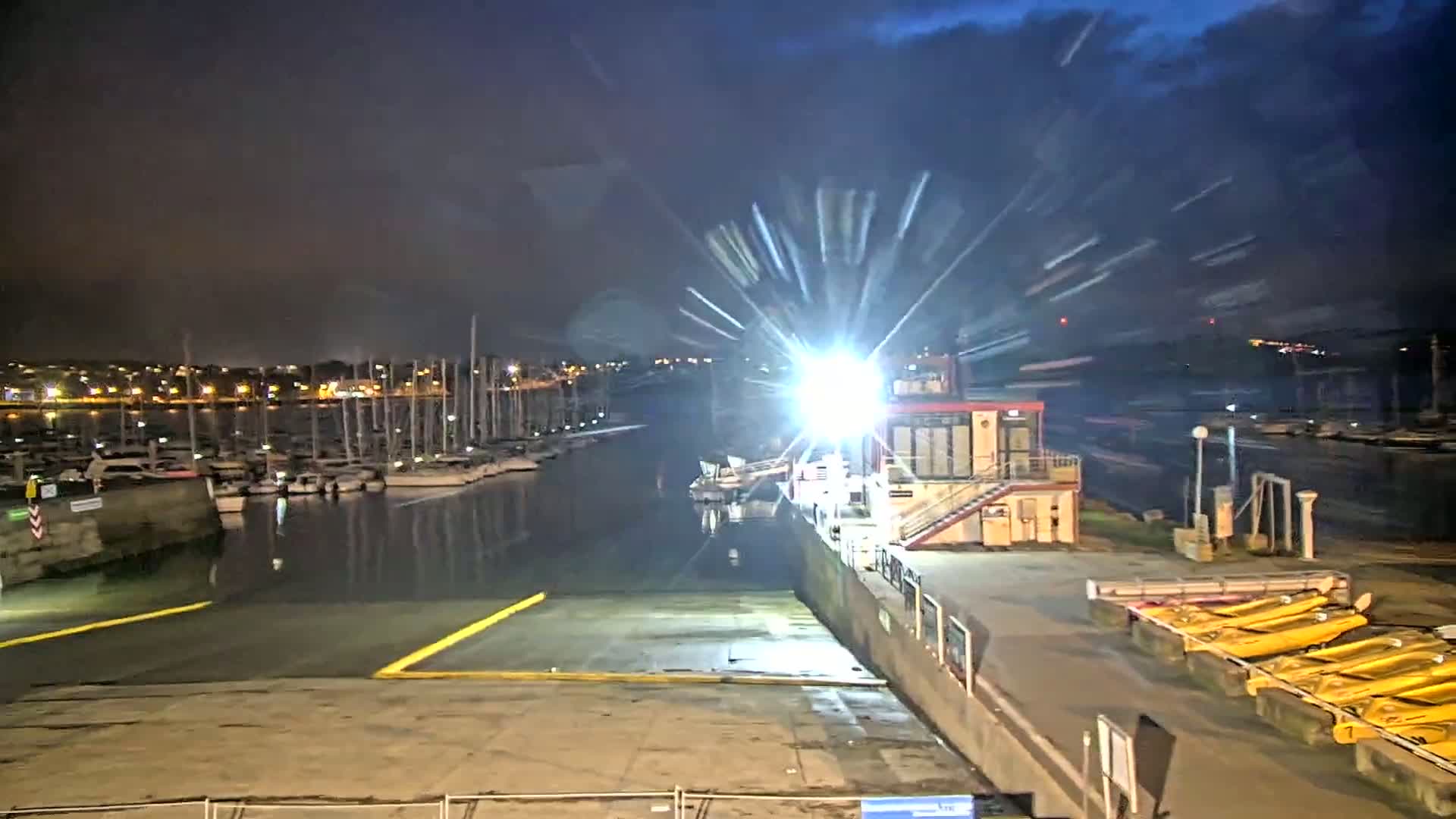 A nighttime view of a marina with numerous boats docked, illuminated by bright lights, under a partly cloudy sky.