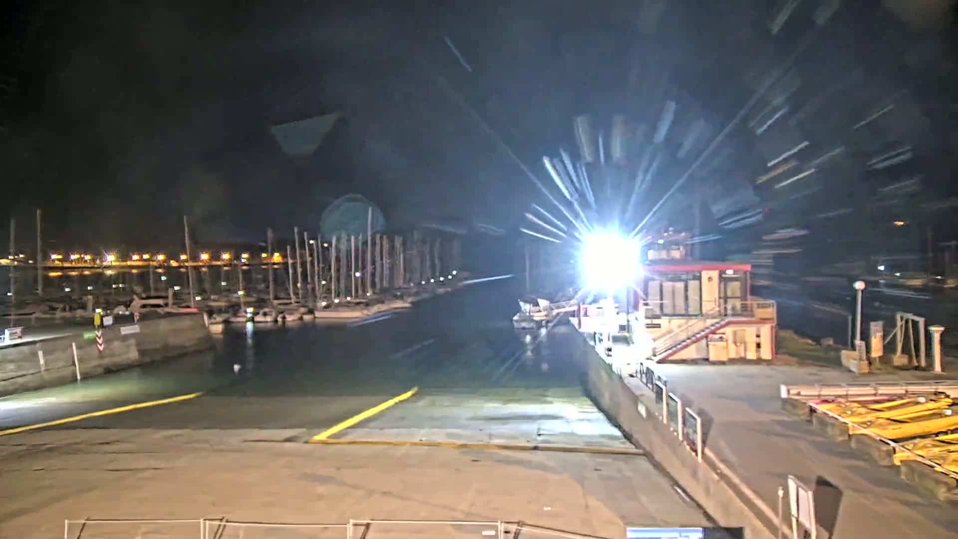 A brightly lit marina at night shows a small building and several docked boats, with a long, yellow-lined dock extending into the water.