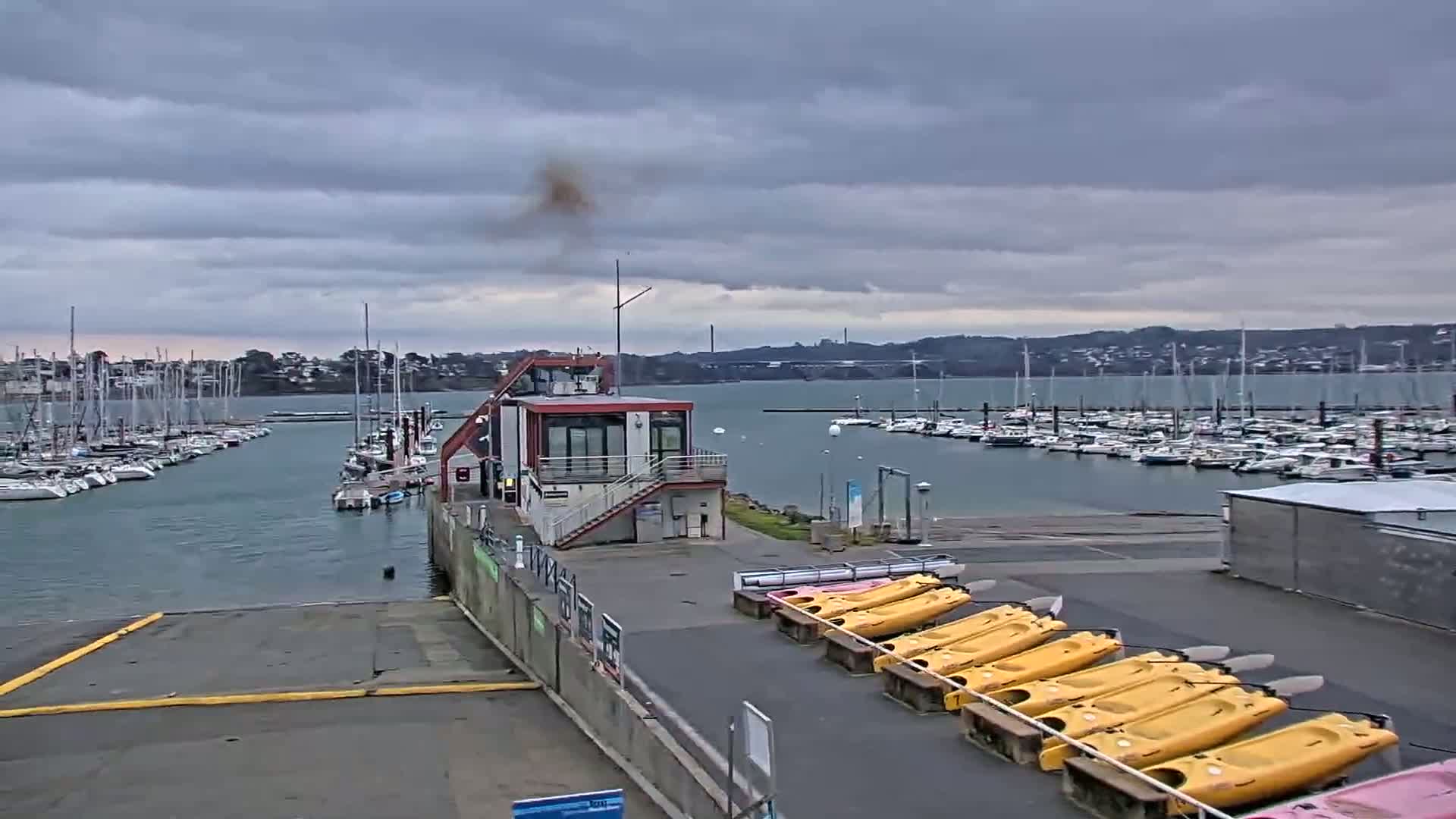 A marina filled with sailboats under a cloudy sky, with several kayaks lined up on the shore near a small building.