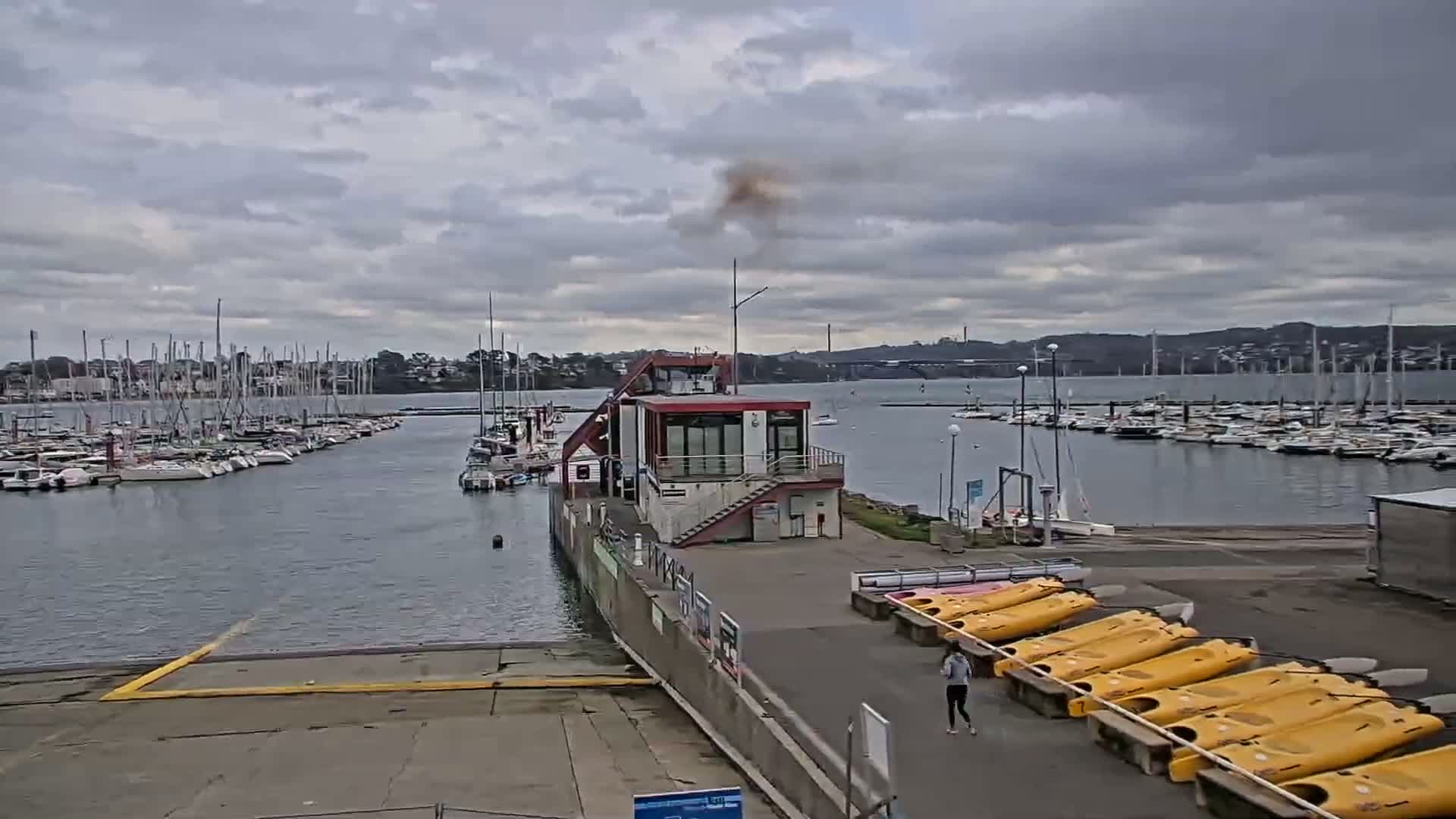 A marina with numerous sailboats and a small building, under a cloudy sky, with a person walking near rows of yellow kayaks.