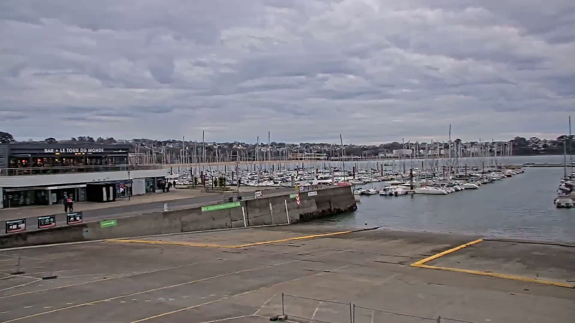 A marina filled with sailboats is seen under a cloudy sky, with a building and paved area in the foreground.