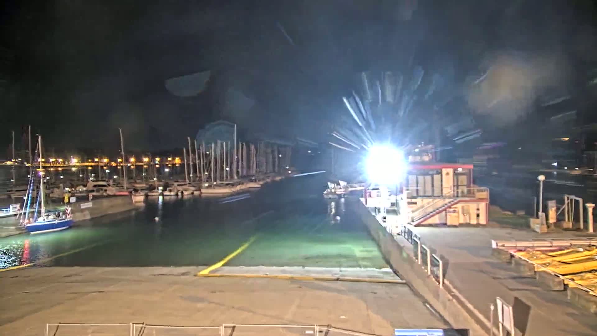 A brightly lit marina at night shows numerous sailboats docked, with a small building and a boat launch area visible.