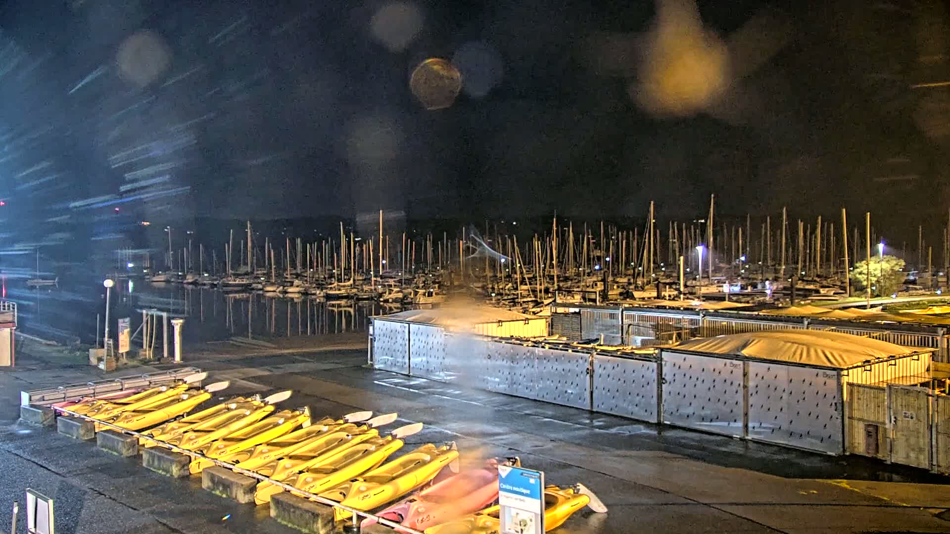 A nighttime view of a marina with numerous sailboats docked, a row of kayaks, and rain falling.