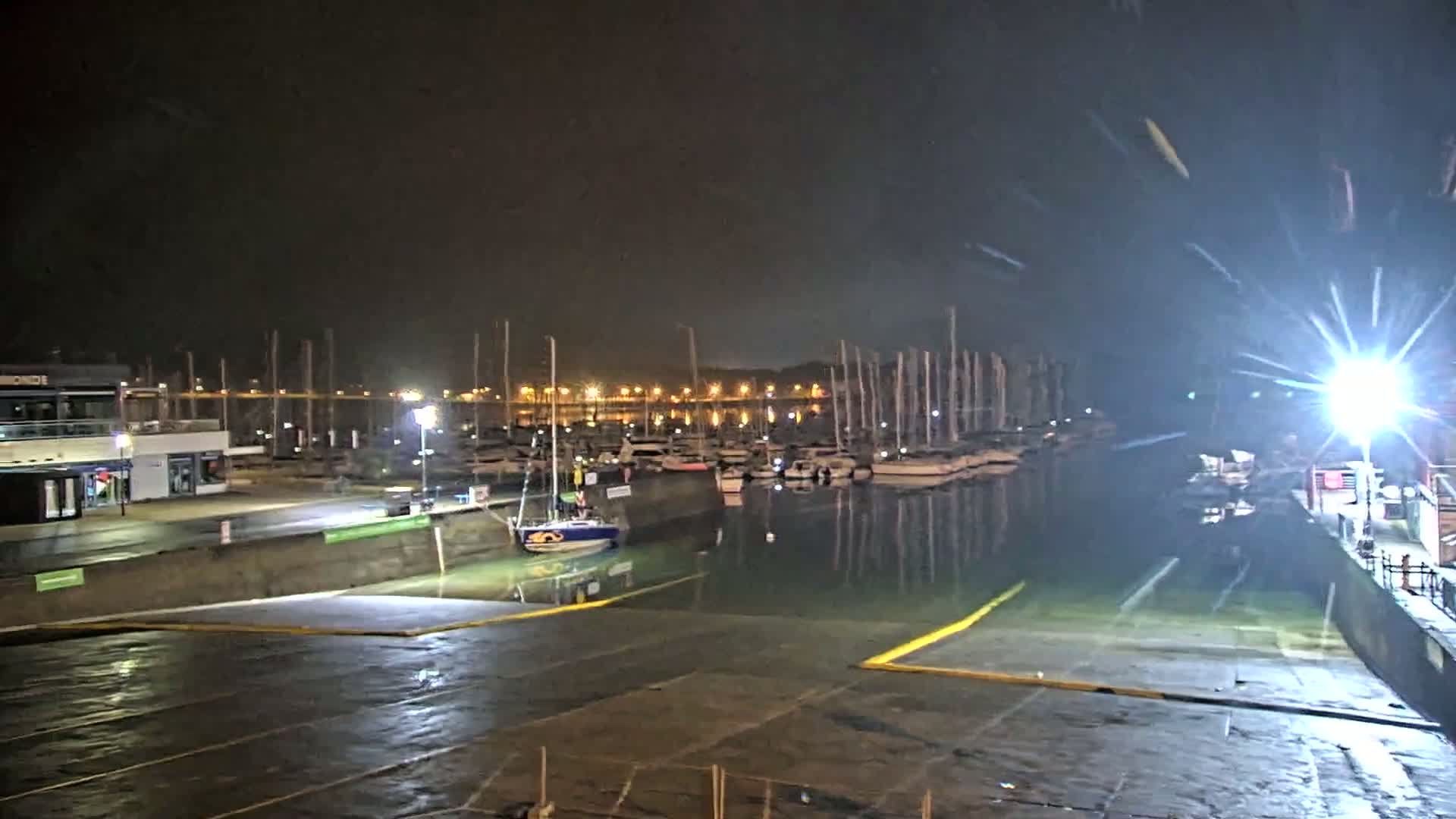 A nighttime view of a marina with numerous boats docked, illuminated by bright lights, under a dark, possibly slightly overcast sky.