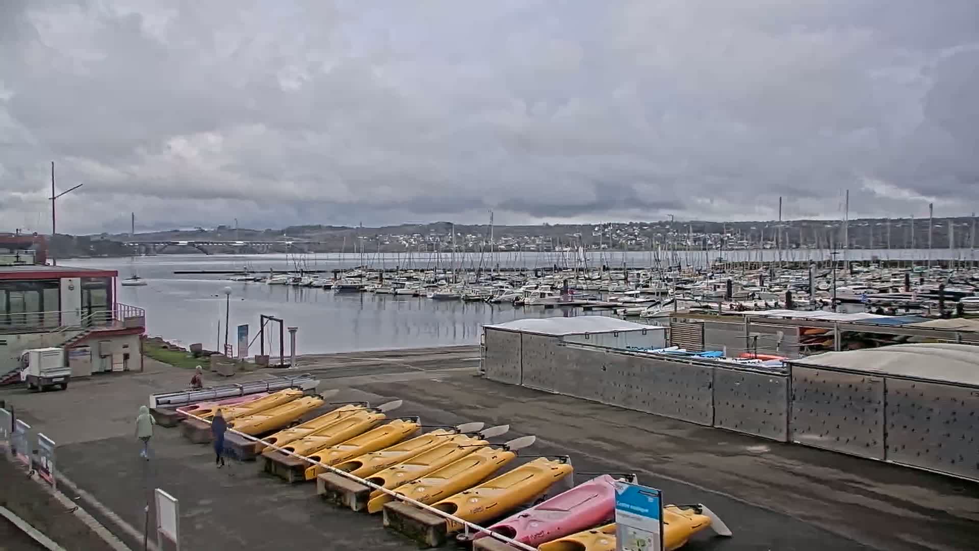 A marina filled with sailboats under a cloudy sky, with kayaks lined up on the shore next to a building.