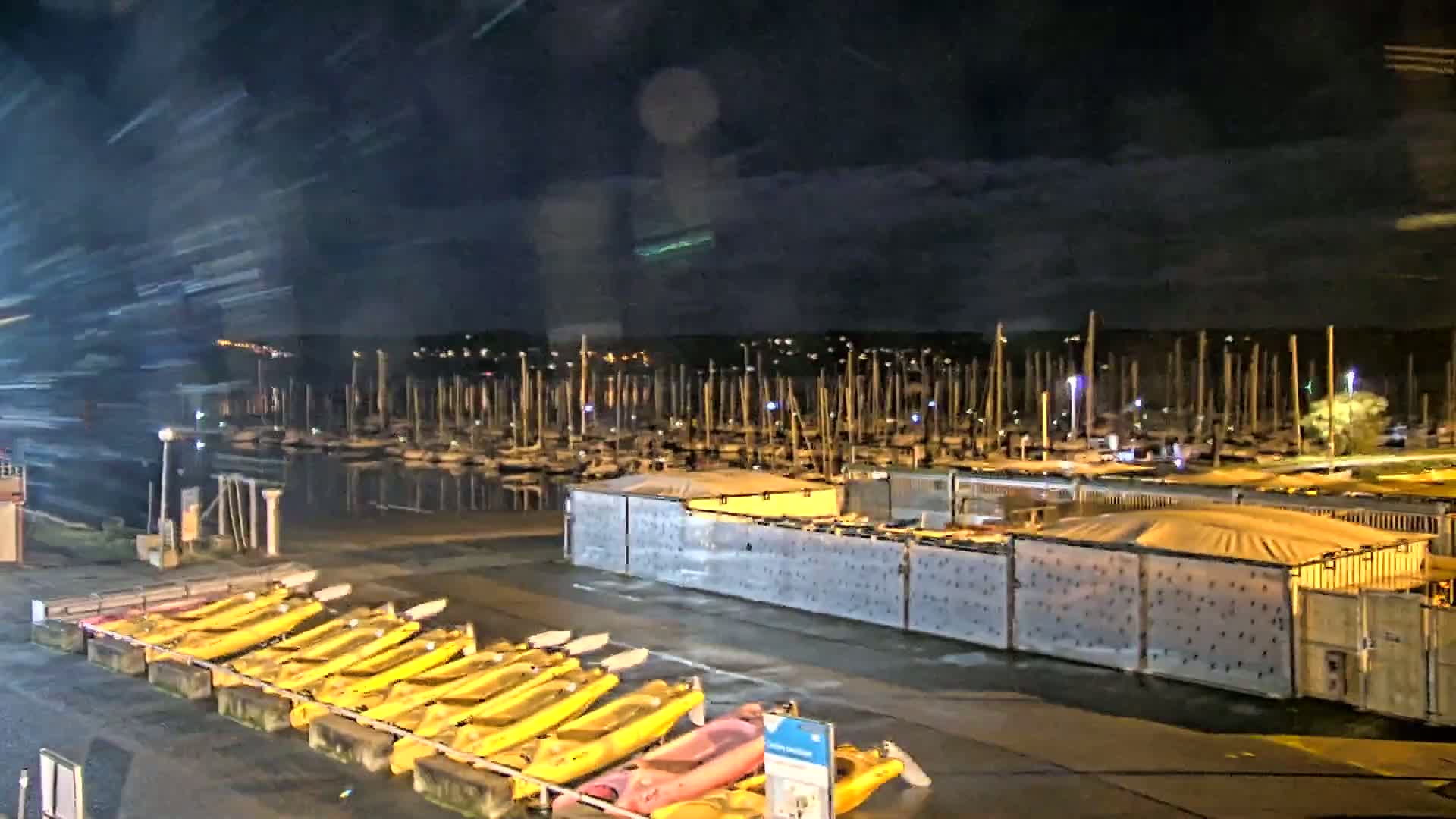 A nighttime view of a marina with numerous boats docked, and rows of yellow kayaks stored on the shore under a dark, possibly overcast sky.