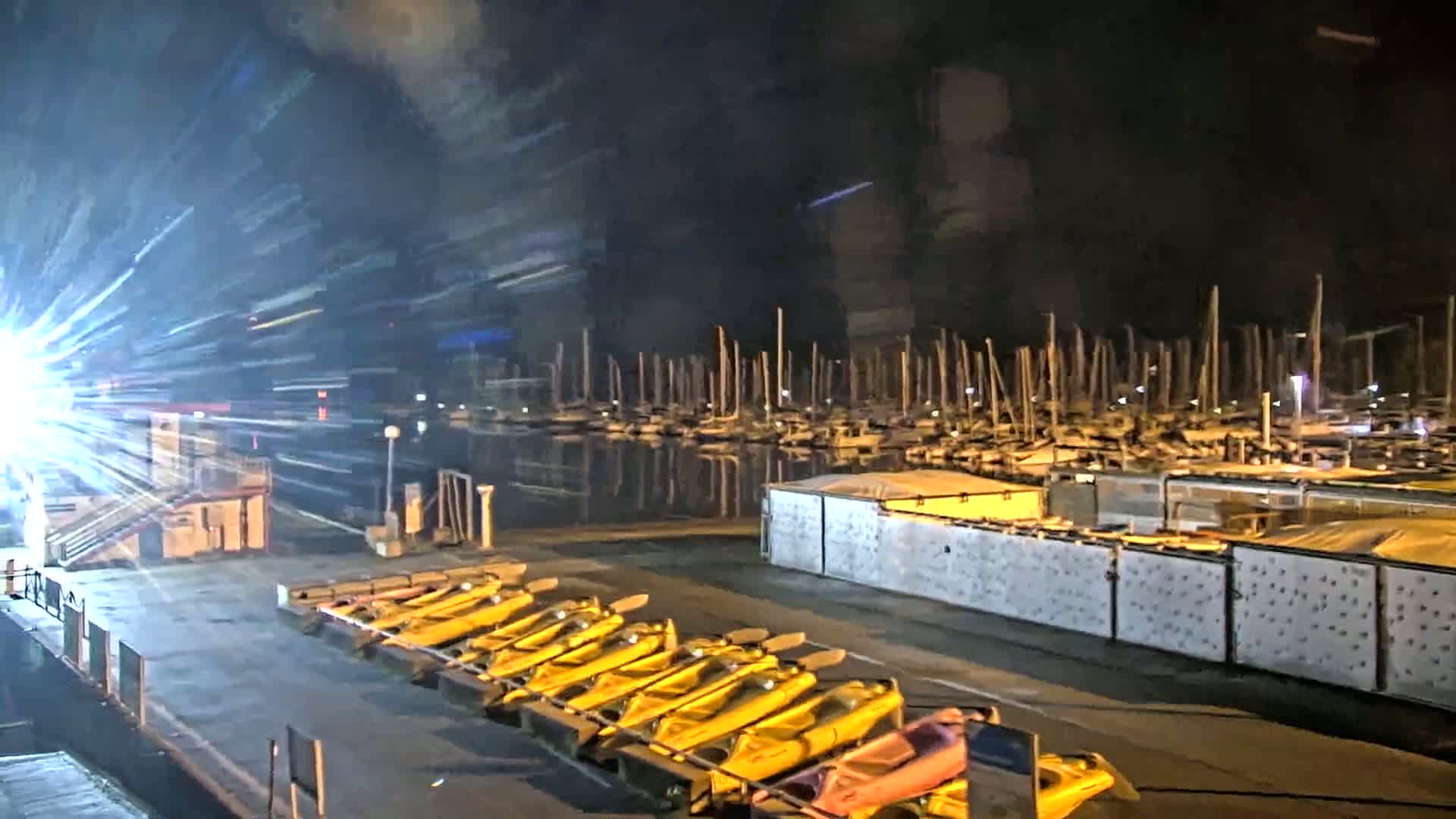 At night, a dock with numerous yellow watercraft is illuminated, adjacent to a marina filled with moored sailboats under a dark sky.