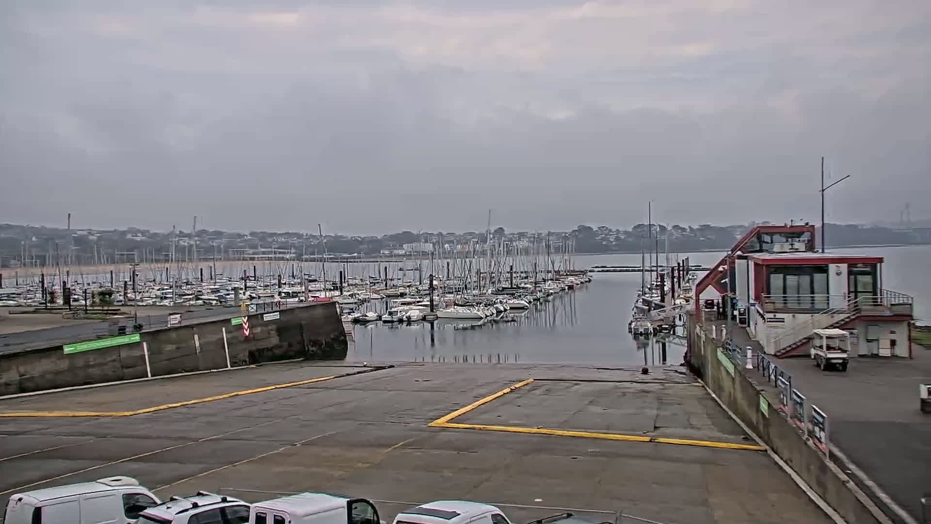 A marina filled with sailboats is seen under an overcast sky, with a boat ramp and several vehicles visible in the foreground.