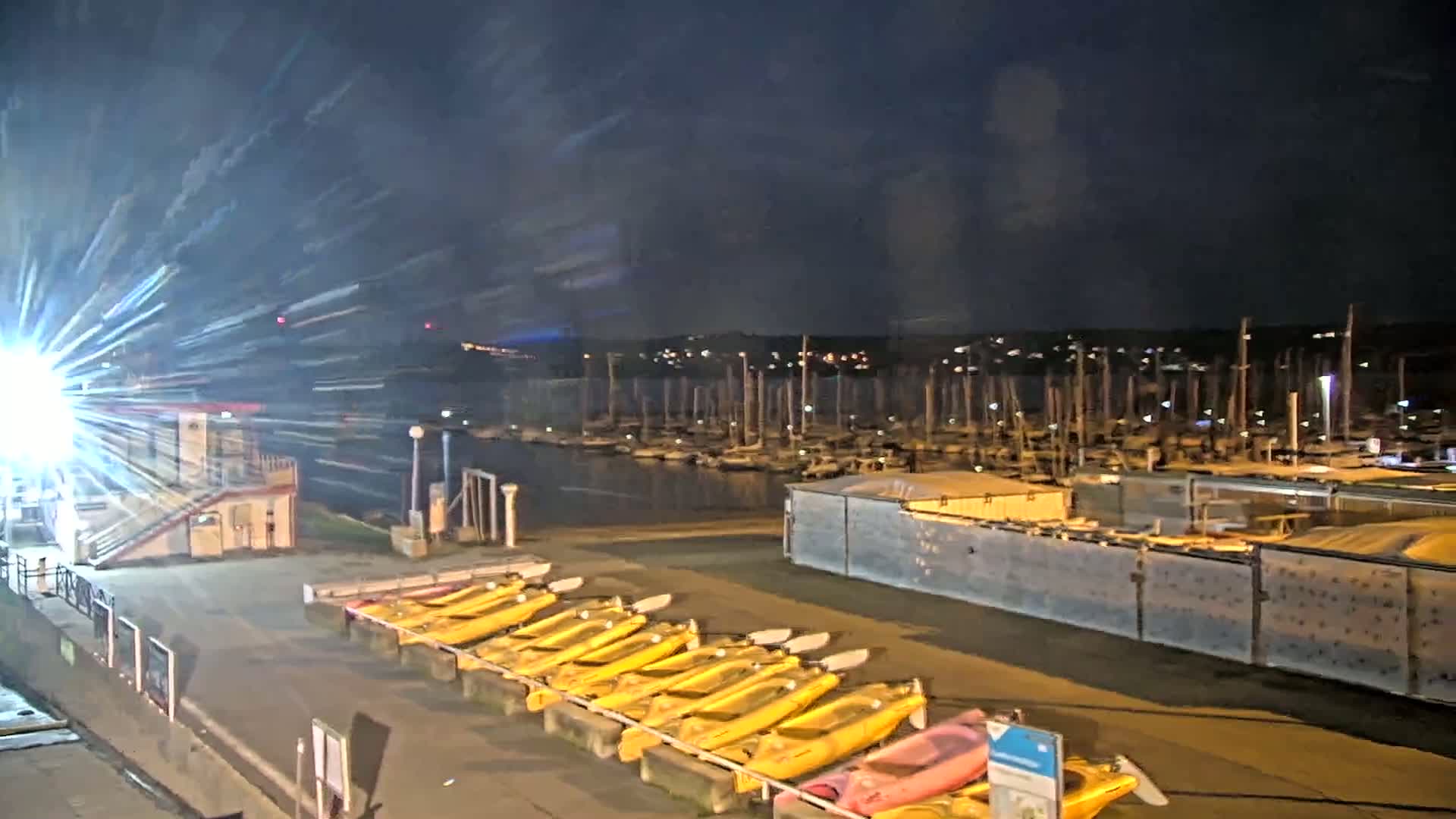 At night, a marina filled with boats is visible beyond a row of yellow kayaks lined up on a dock under a dark sky.