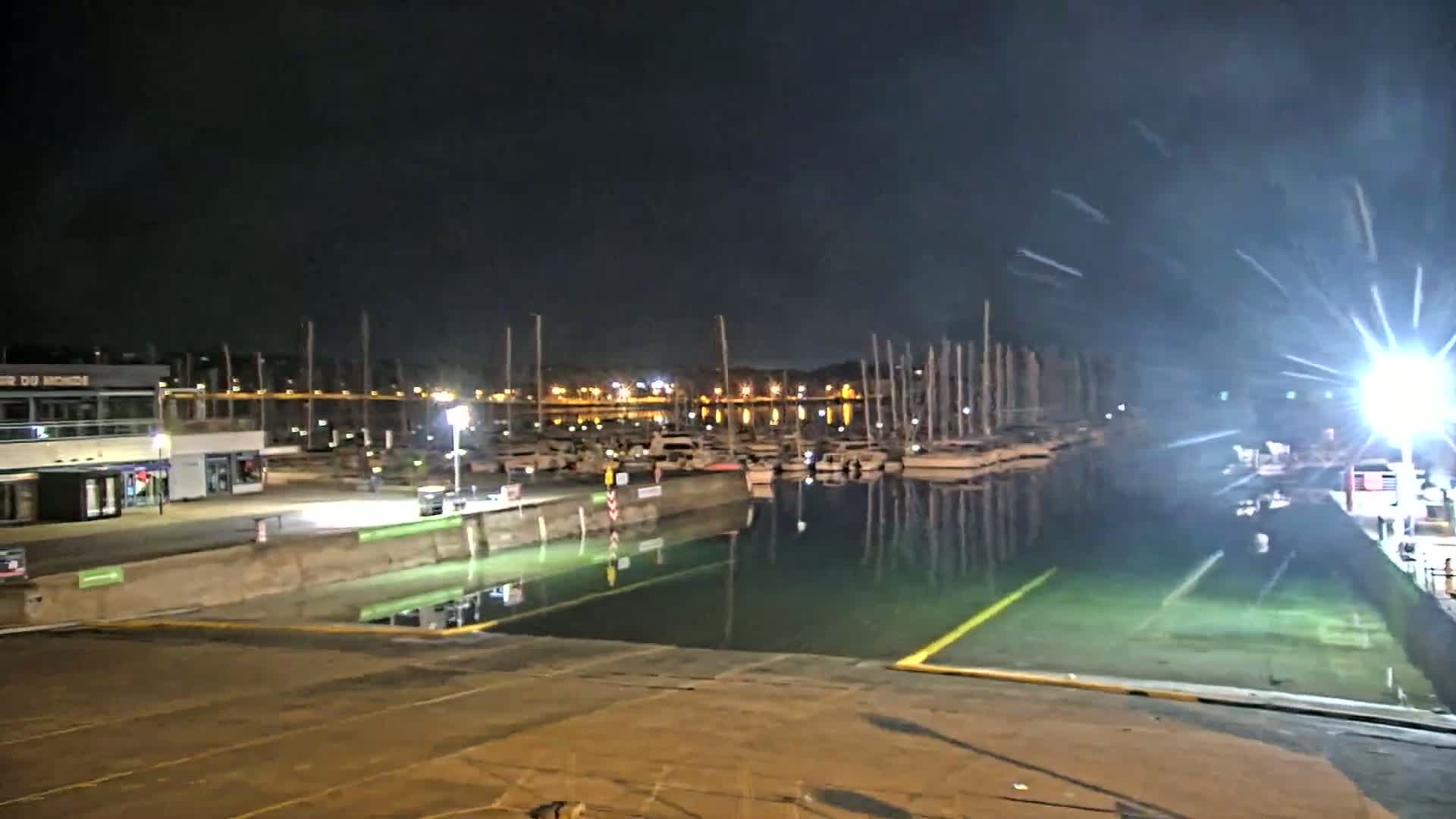 A nighttime view of a marina with numerous boats docked, illuminated by artificial lights, under a dark sky.