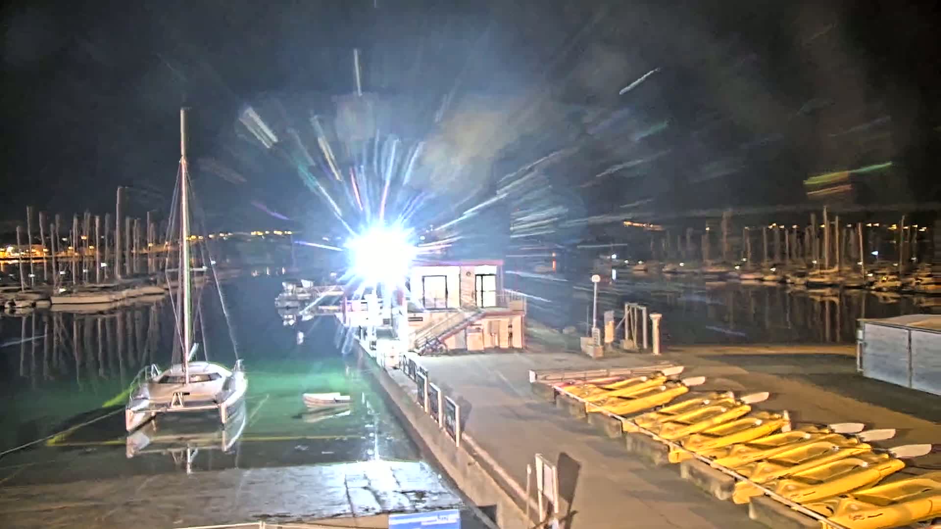 At night, a marina with docked sailboats and kayaks is brightly lit, with a bright light creating starburst streaks in the image.