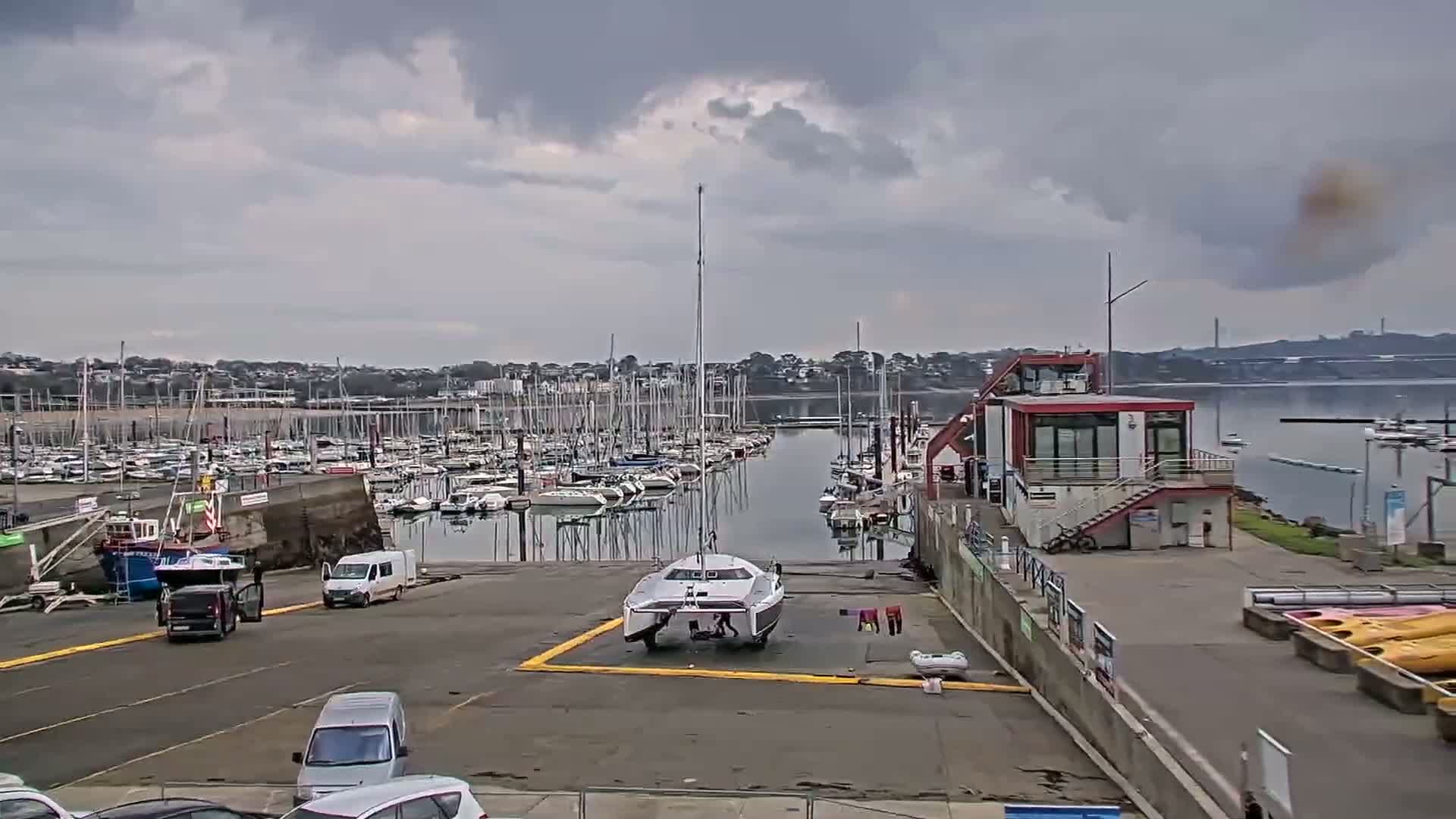 A marina filled with sailboats under a cloudy sky, with a catamaran on a launch ramp and several vehicles parked nearby.