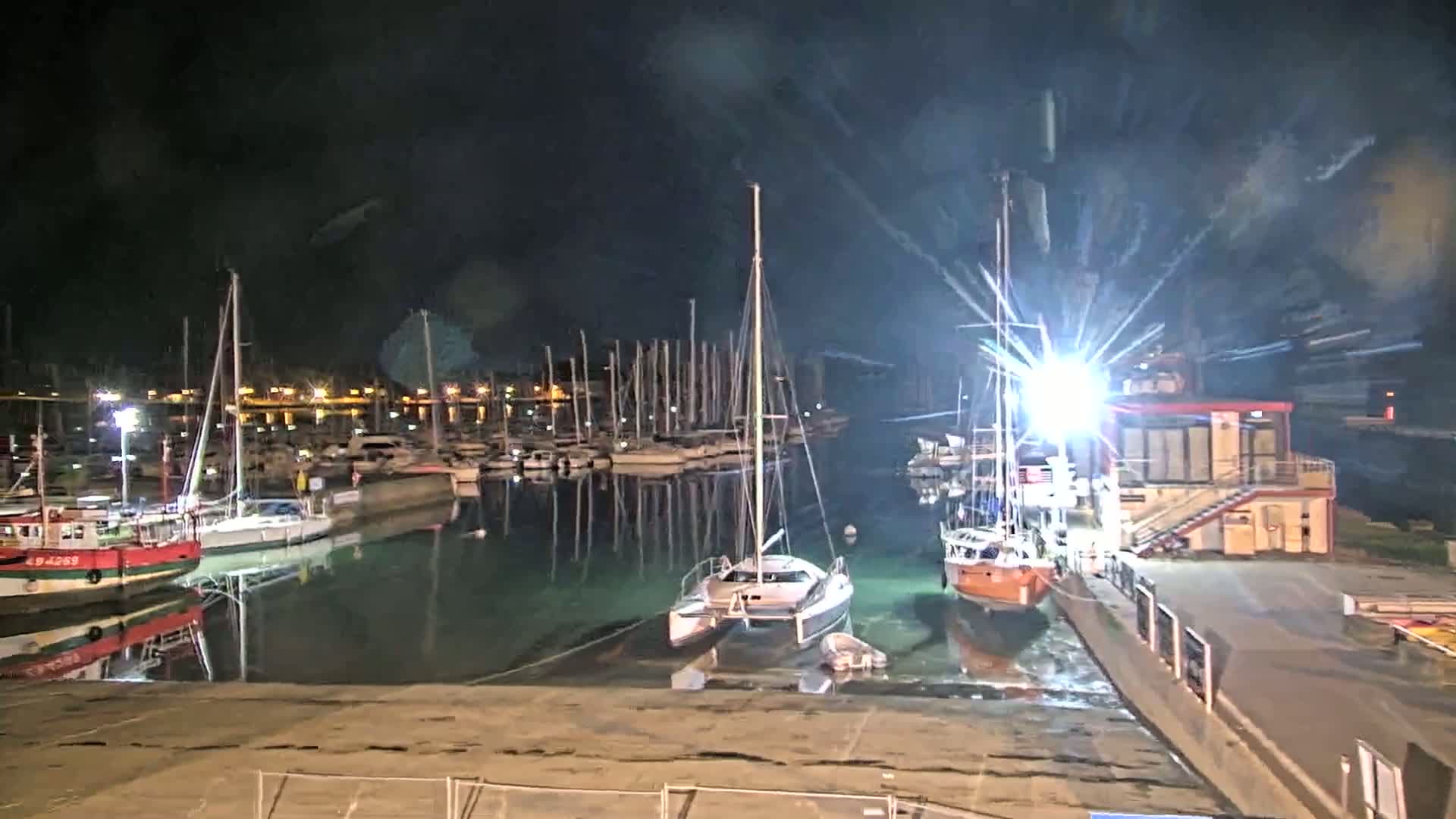 Many sailboats and motorboats are docked at a marina at night under a dark sky.
