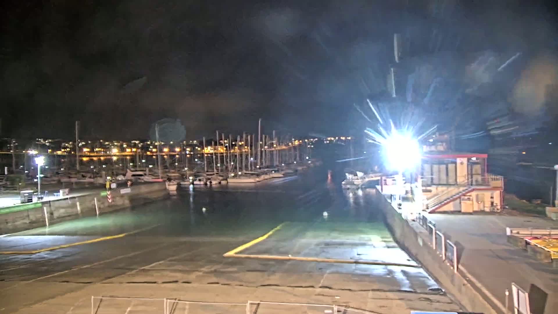 At night, a marina filled with docked sailboats is brightly lit, with a floodlight creating a starburst effect near a small building.
