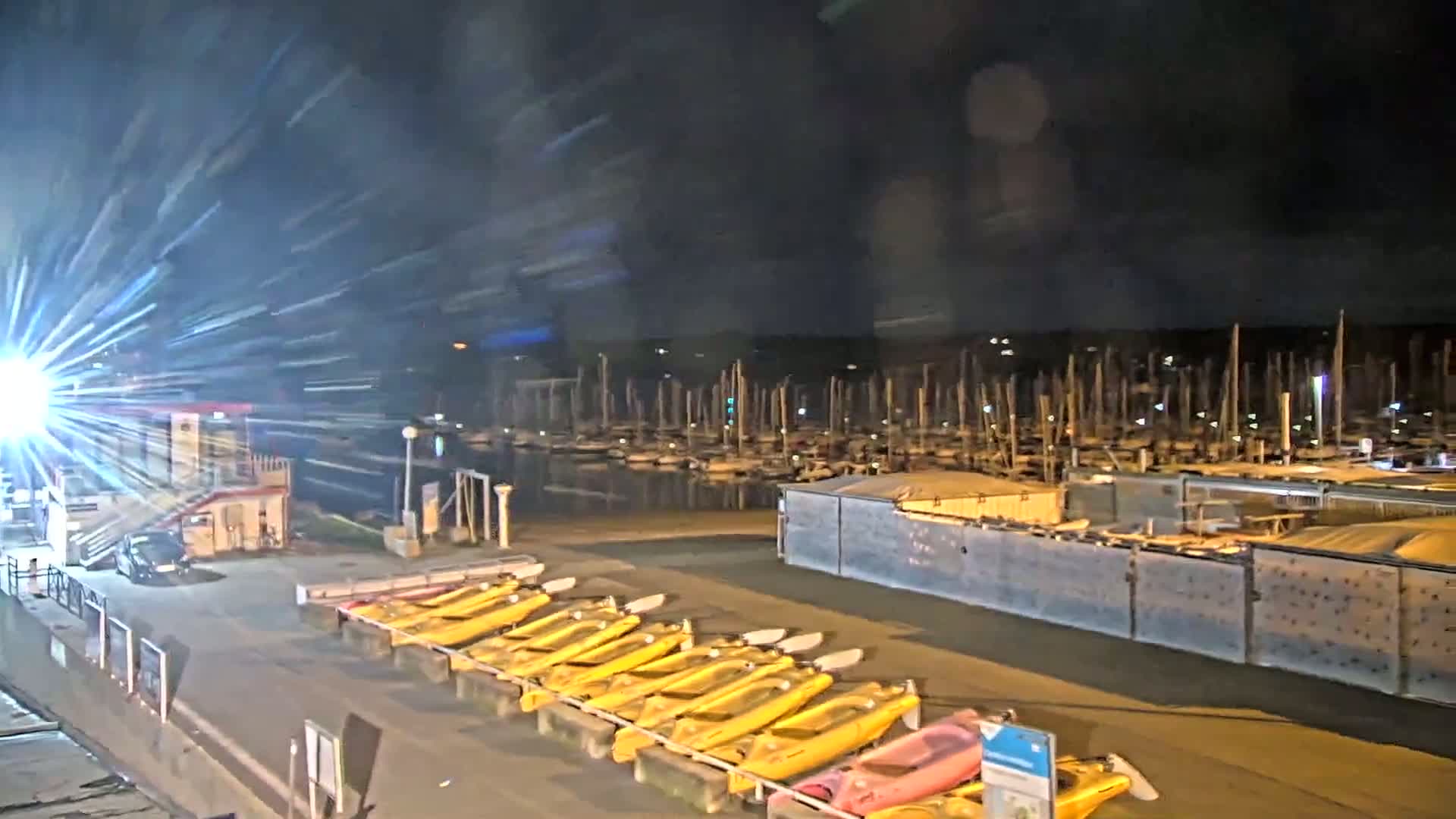 At night, a marina filled with boats is visible beyond a row of yellow kayaks lined up near a building with a bright light shining outward.