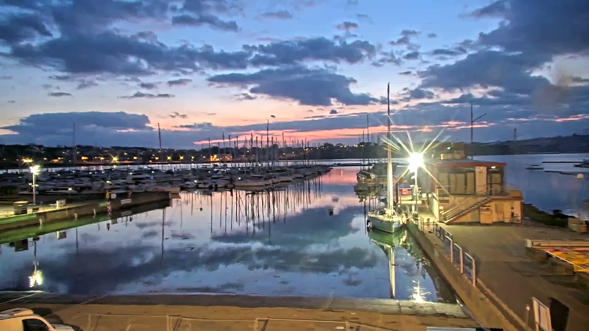 Many sailboats are docked in a marina at sunset under a partly cloudy sky.
