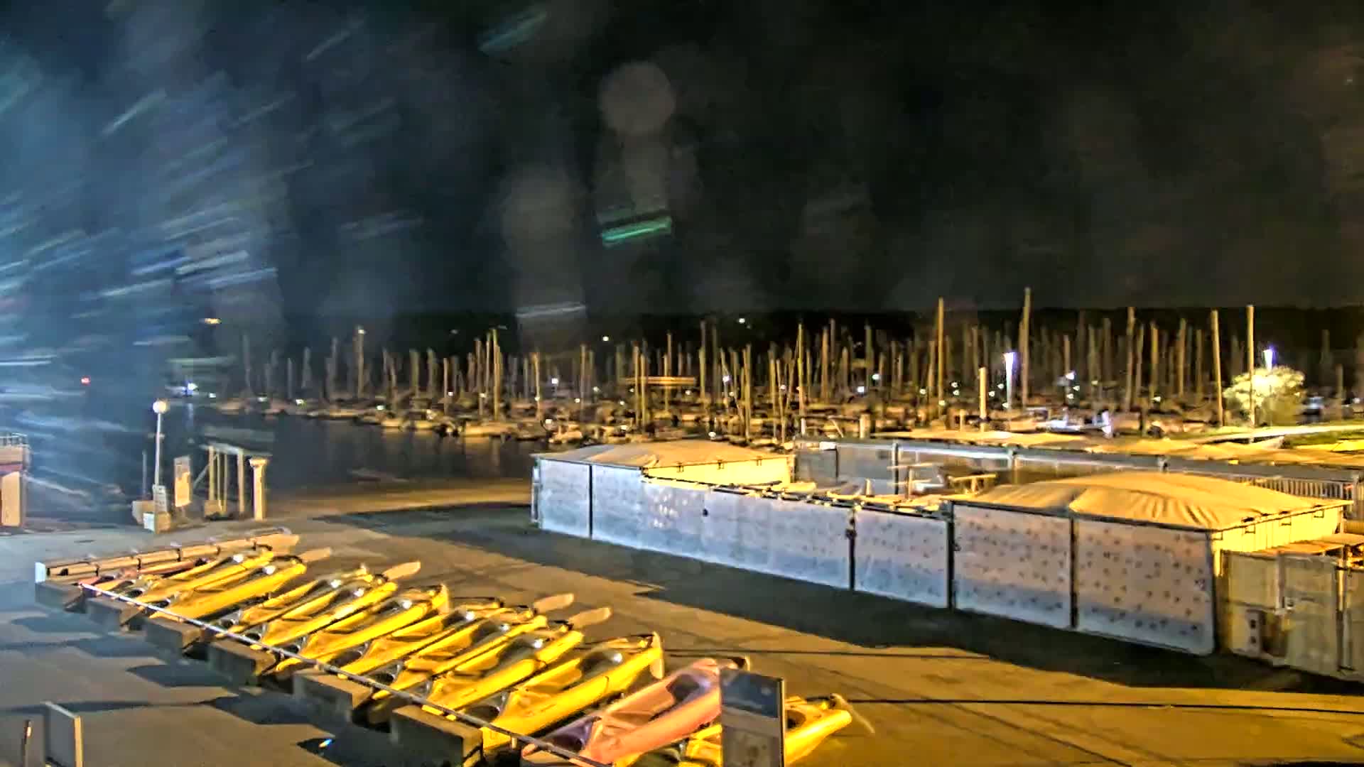 A nighttime view of a marina with numerous boats moored, a row of yellow kayaks on a rack, and several covered storage containers, with motion blur suggesting movement in the foreground.