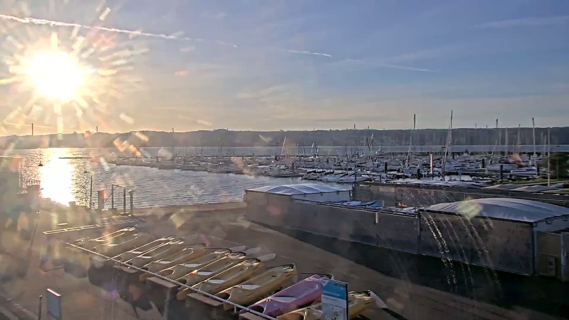 A bright sunrise shines over a calm harbor filled with numerous boats and sailboats, with kayaks lined up on a rack in the foreground.