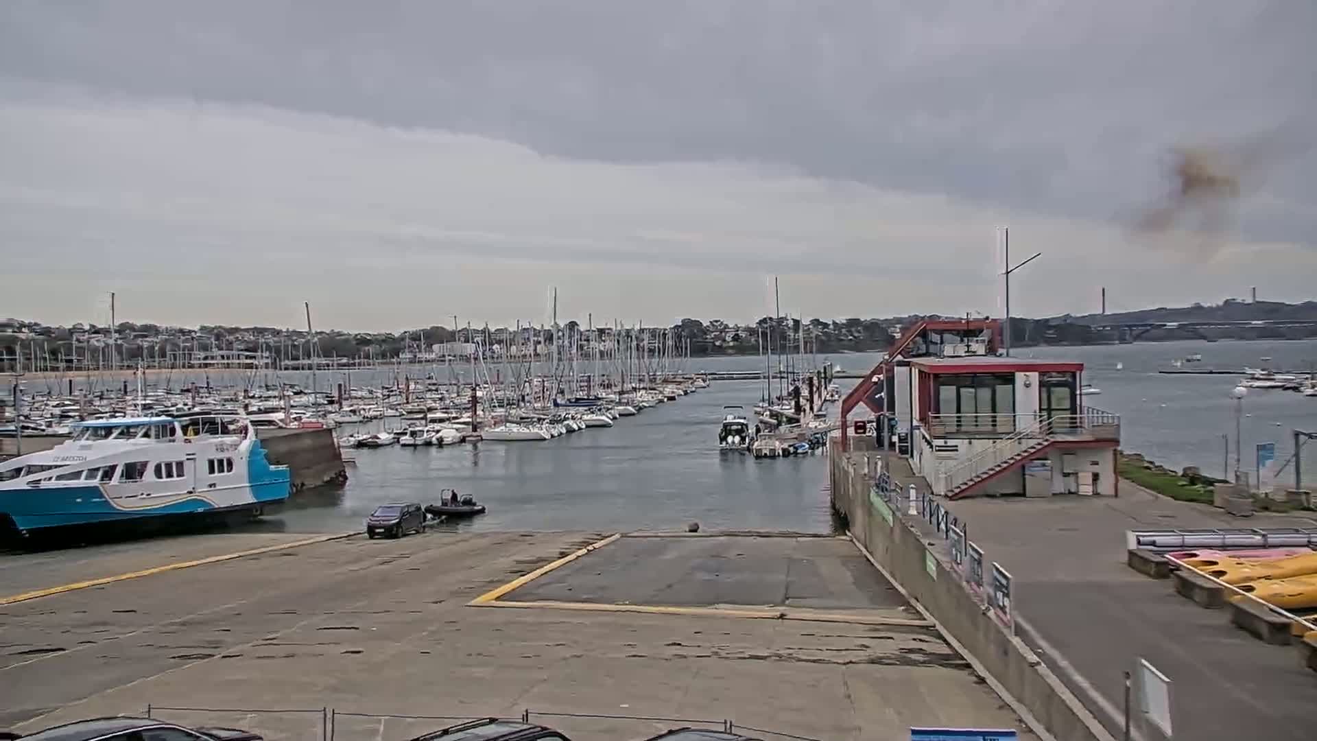 A marina filled with numerous sailboats under a cloudy sky, with a ferry and small building nearby.