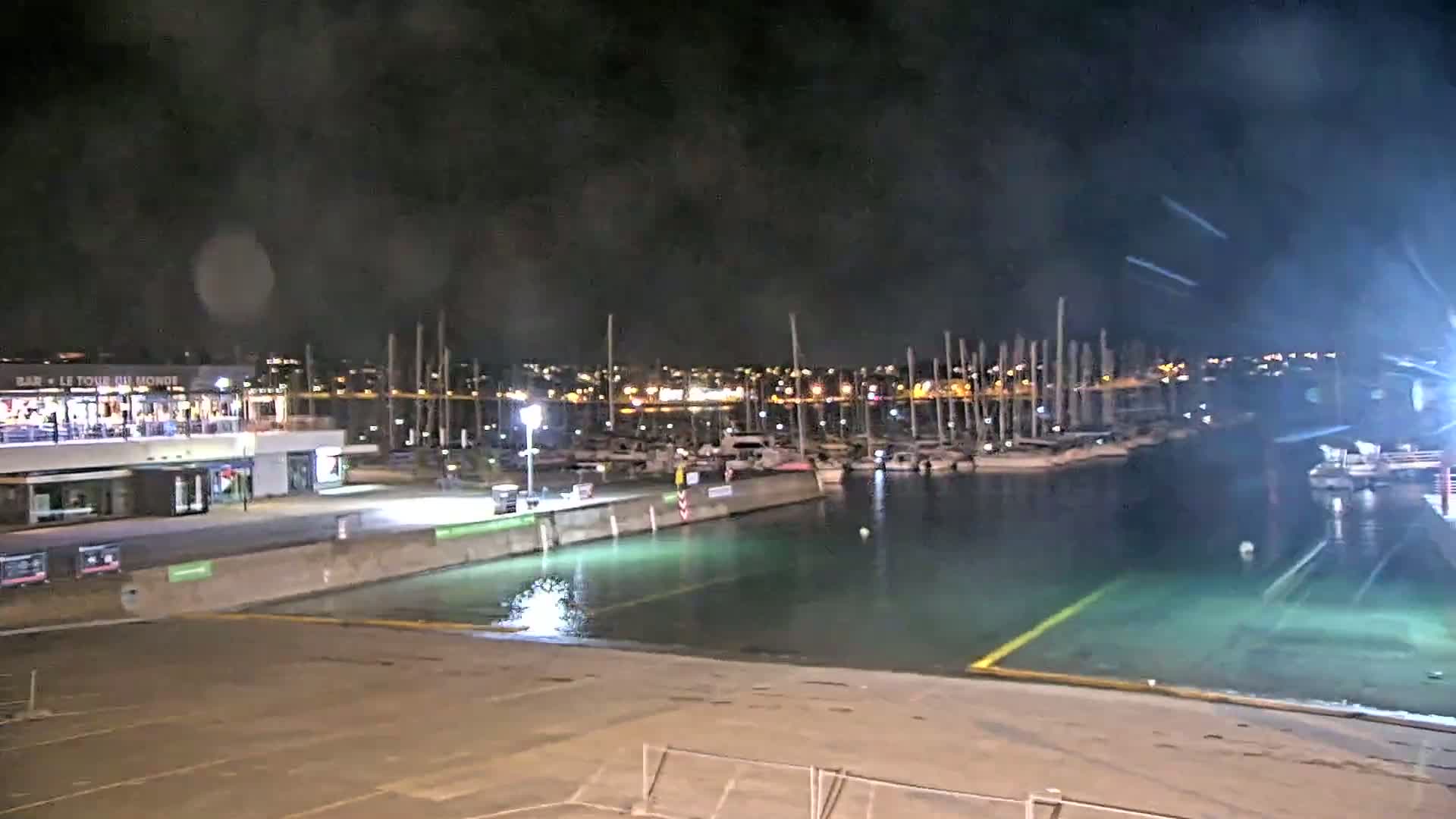 A nighttime view of a marina with many sailboats docked, under a dark, cloudy sky.