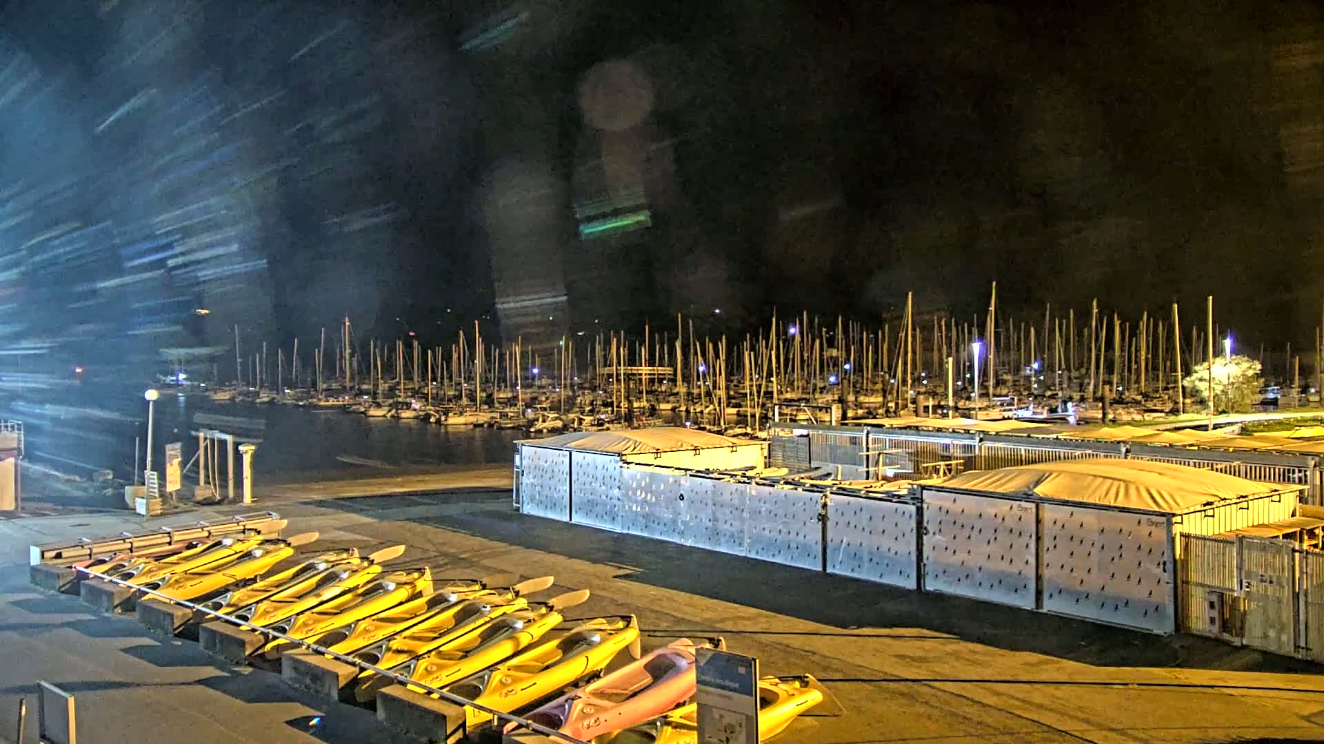 At night, numerous yellow kayaks are lined up on a dock next to a marina filled with sailboats under a dark, possibly stormy sky.