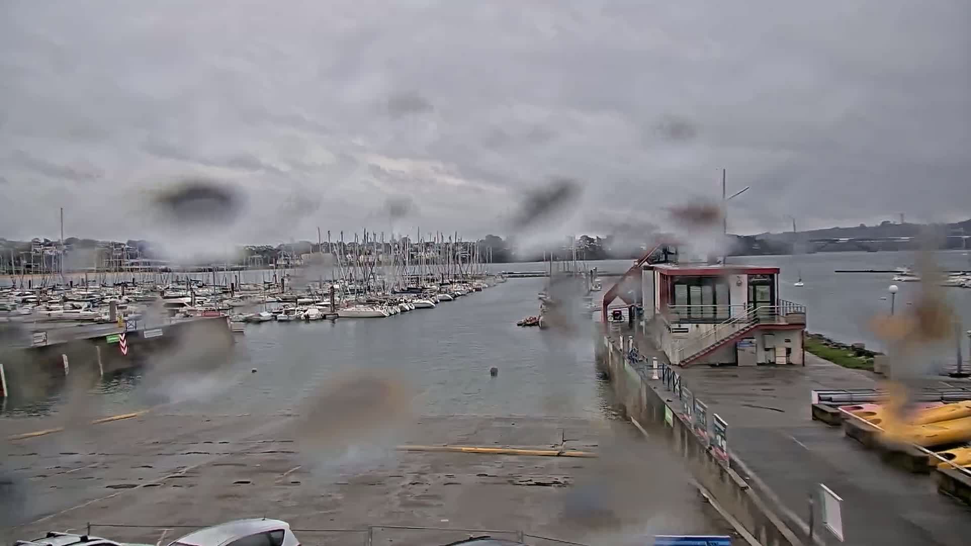 A marina filled with sailboats is seen through rain-streaked glass on a cloudy day.