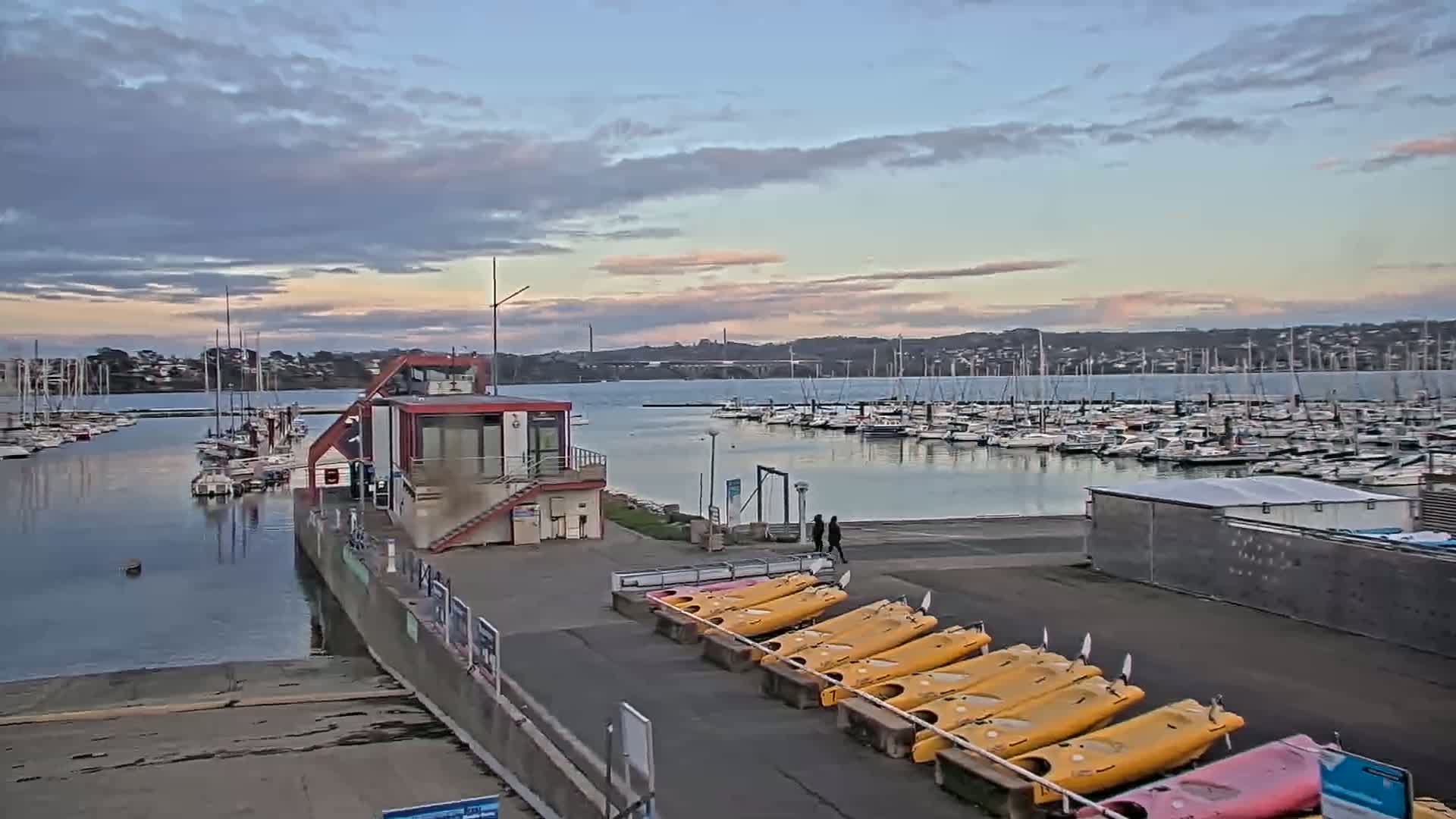 A marina filled with sailboats under a partly cloudy, dusky sky, with kayaks lined up on a dock near a small building.