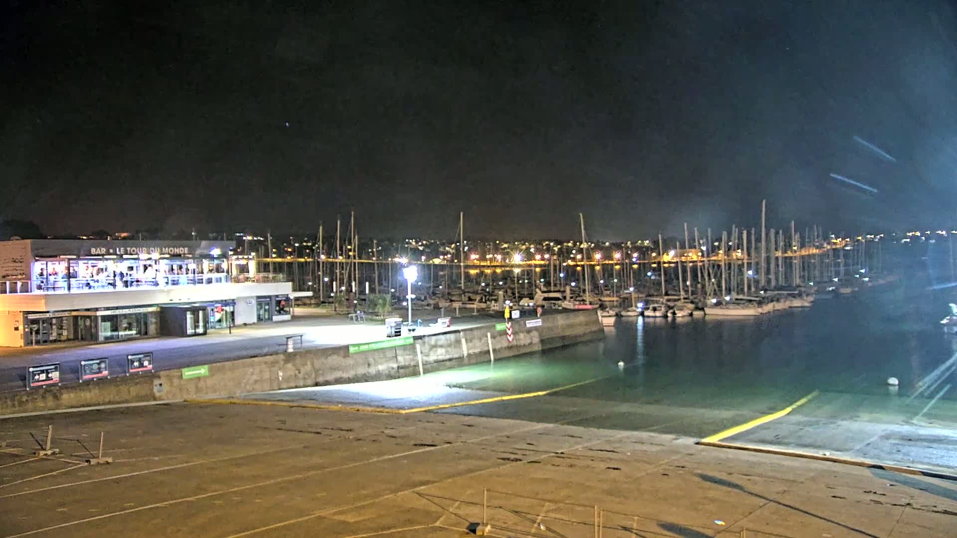 A brightly lit marina at night, filled with docked sailboats, is adjacent to a waterfront building with lights on.