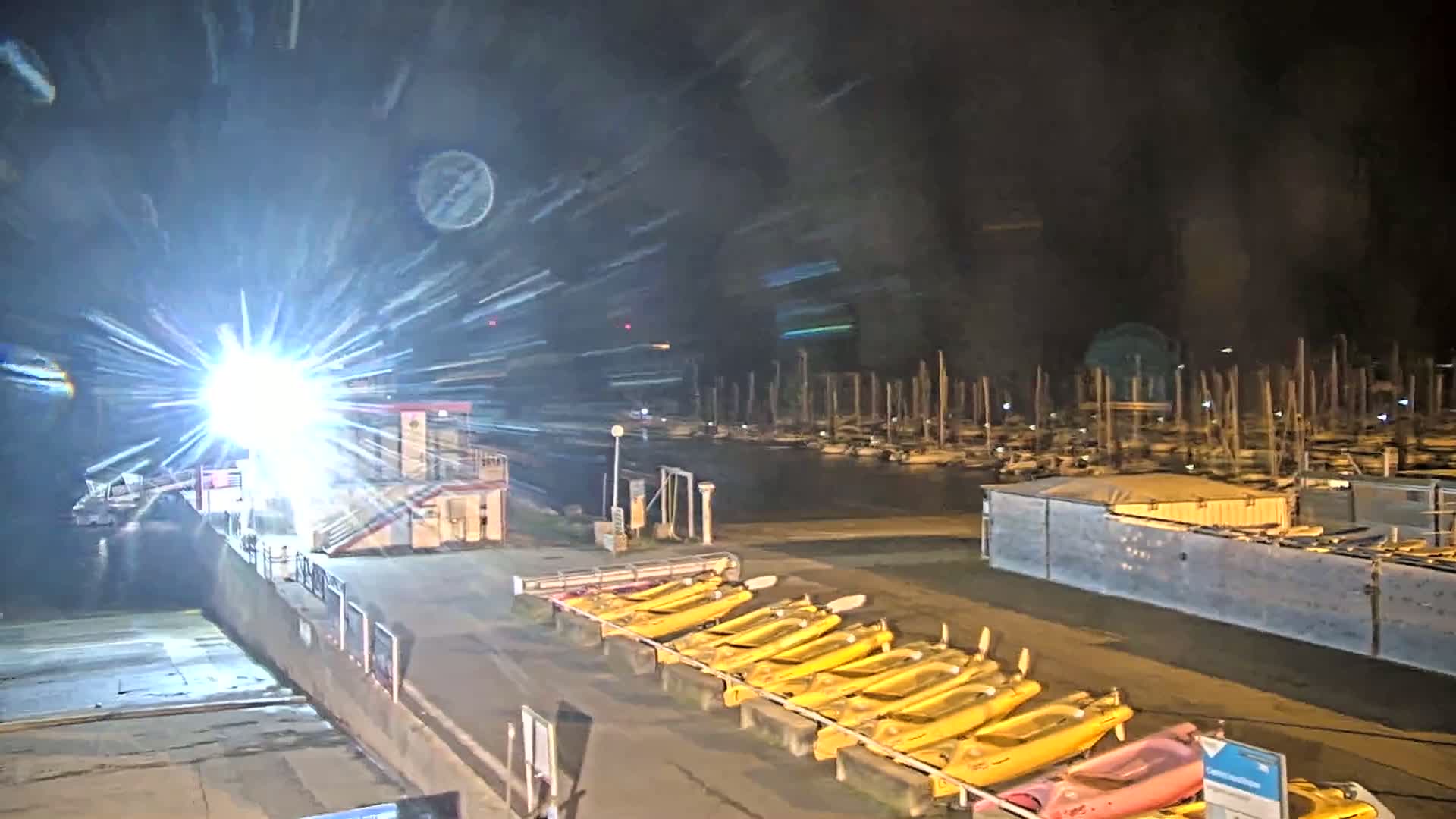 A nighttime view of a marina with many boats moored, a bright light shining, and a row of yellow kayaks lined up on the dock.