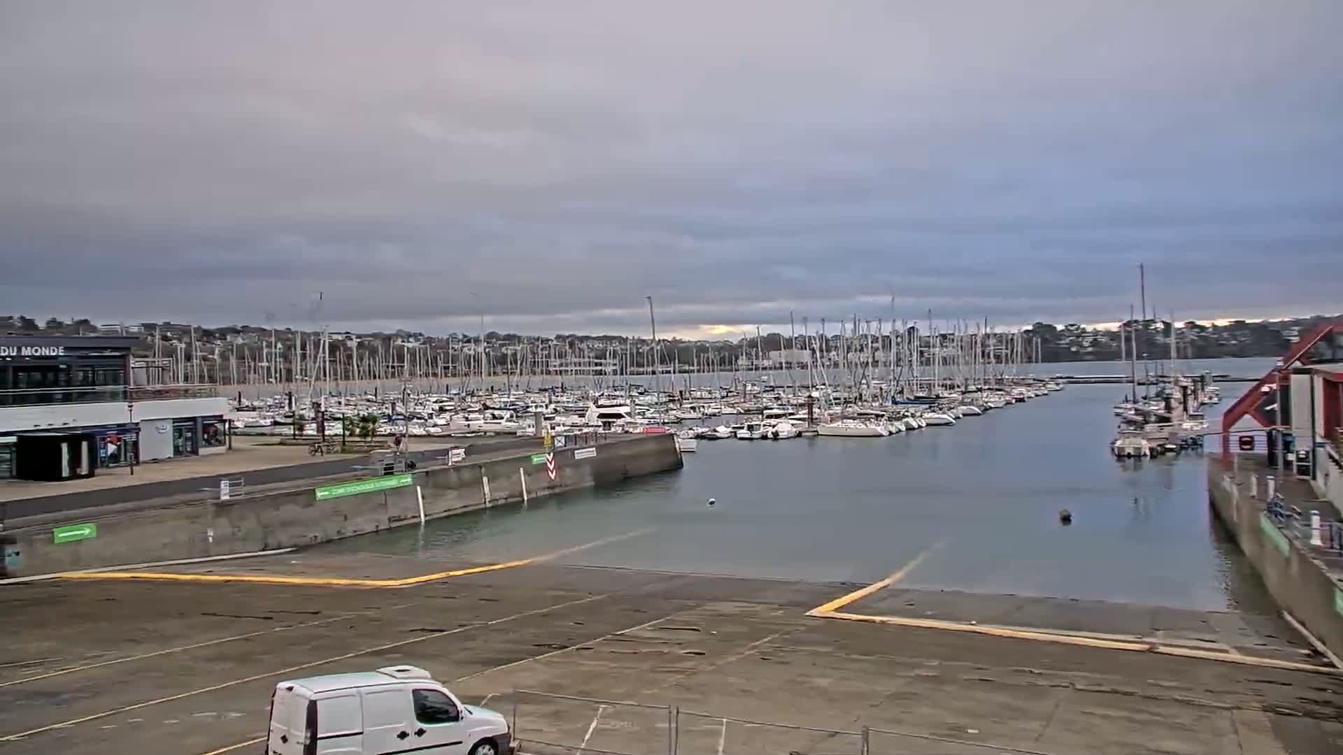 A marina filled with numerous sailboats is viewed from a paved area under an overcast sky.