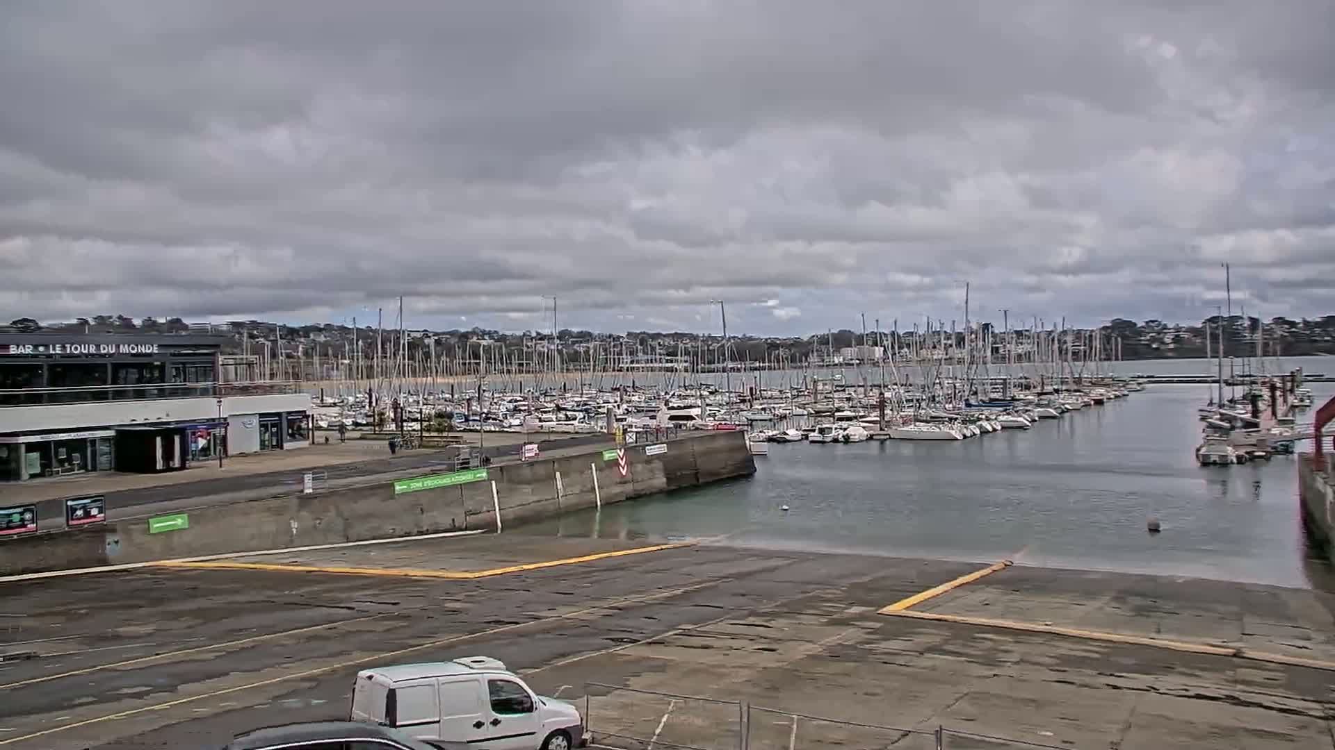 A marina filled with sailboats is seen under an overcast sky, with a paved area in the foreground and a building along the waterfront.