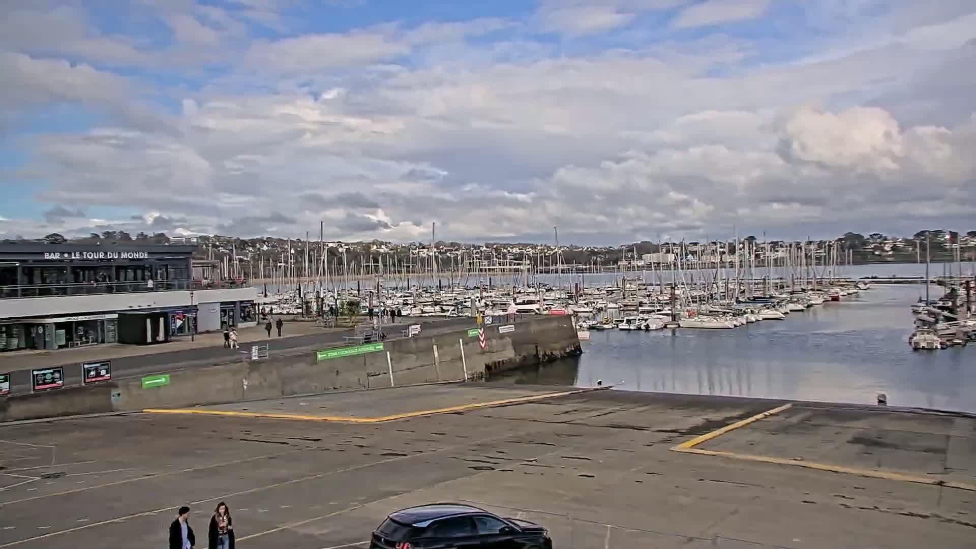A mostly cloudy day reveals a marina filled with sailboats, overlooked by a building and an empty parking lot with a couple walking by.
