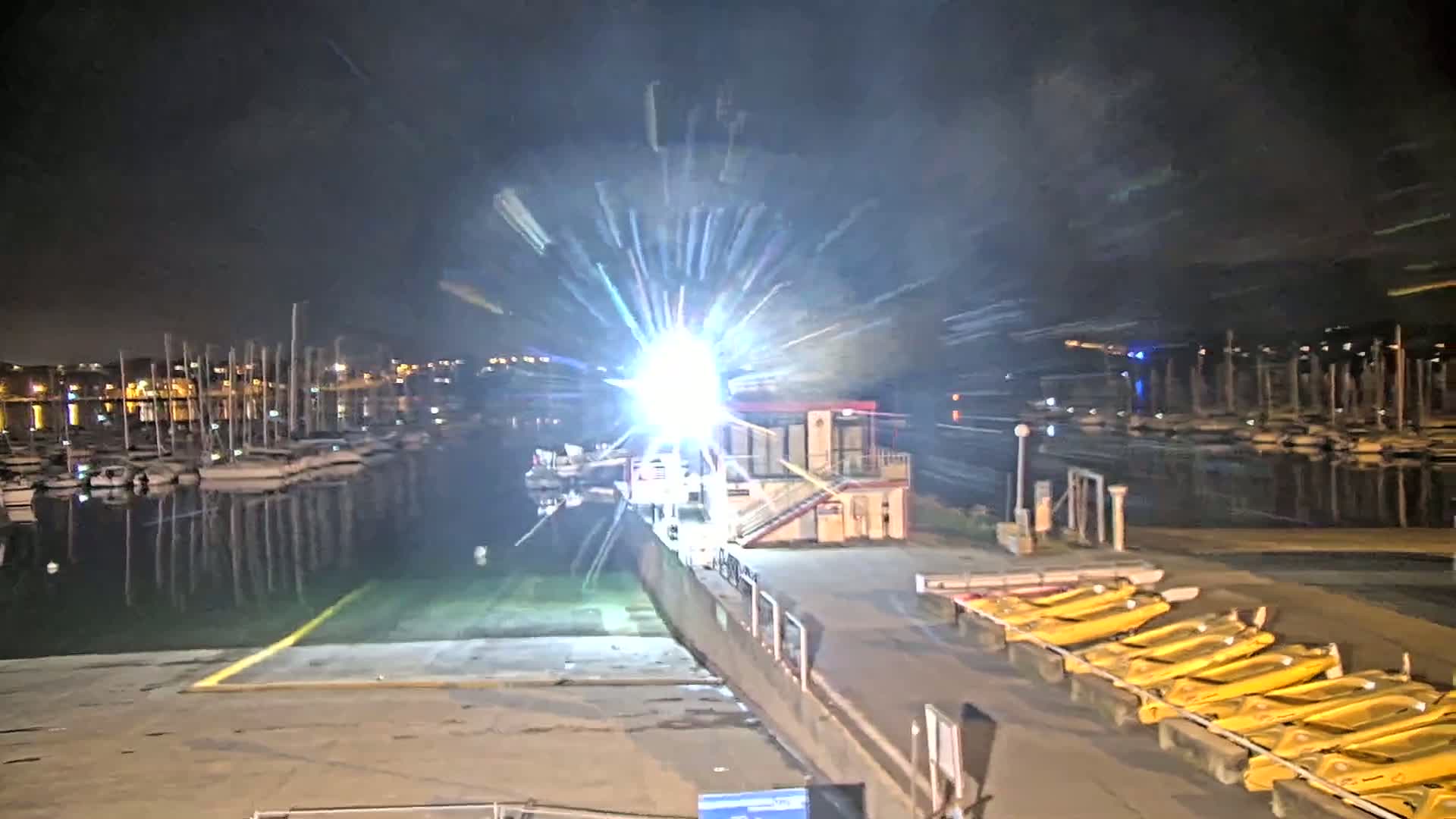 A bright light shines over a marina at night, illuminating numerous boats moored along a dock with yellow kayaks lined up nearby.