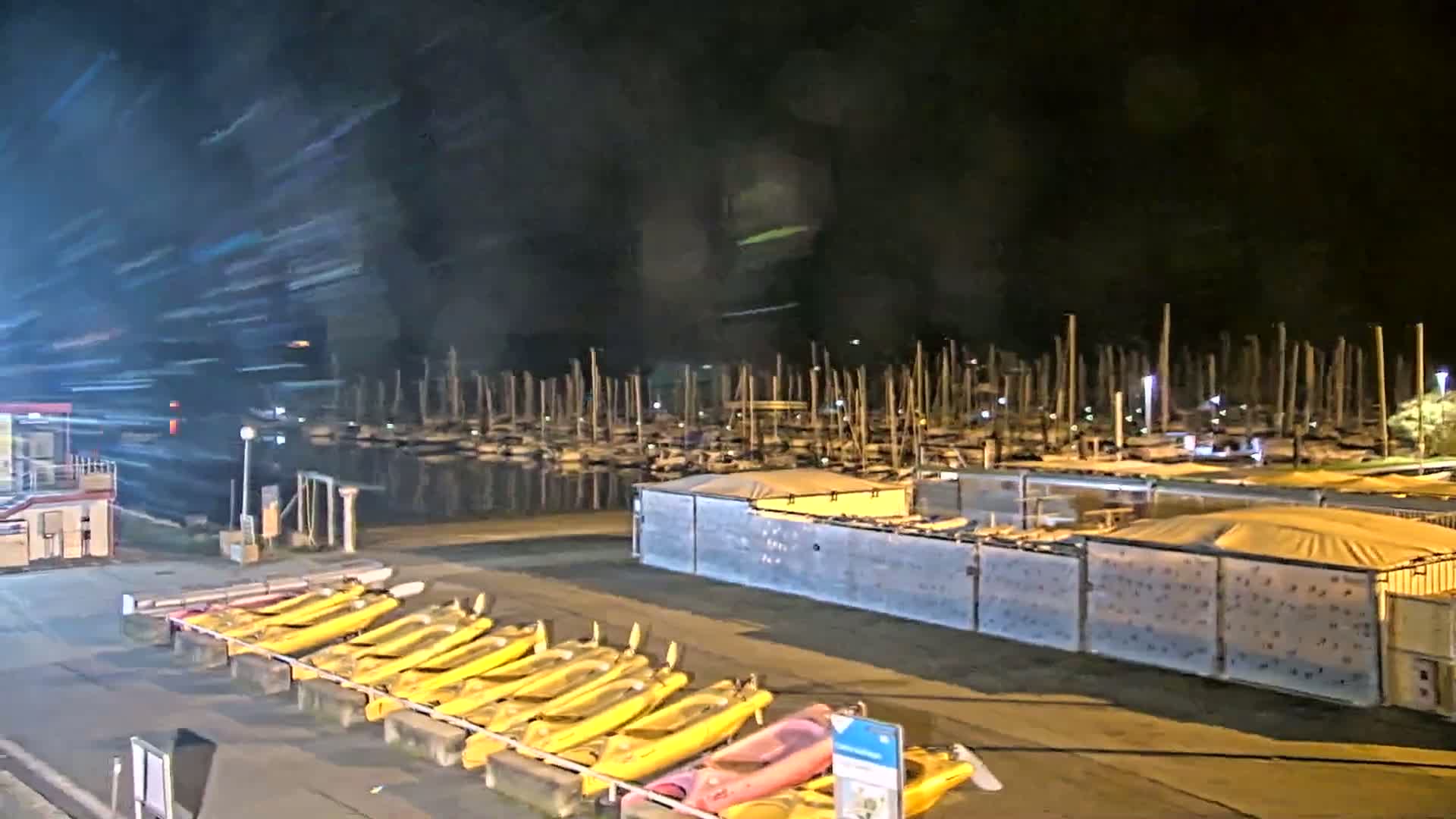 At night, a marina with numerous docked sailboats is visible beyond a row of yellow kayaks lined up along a paved area.