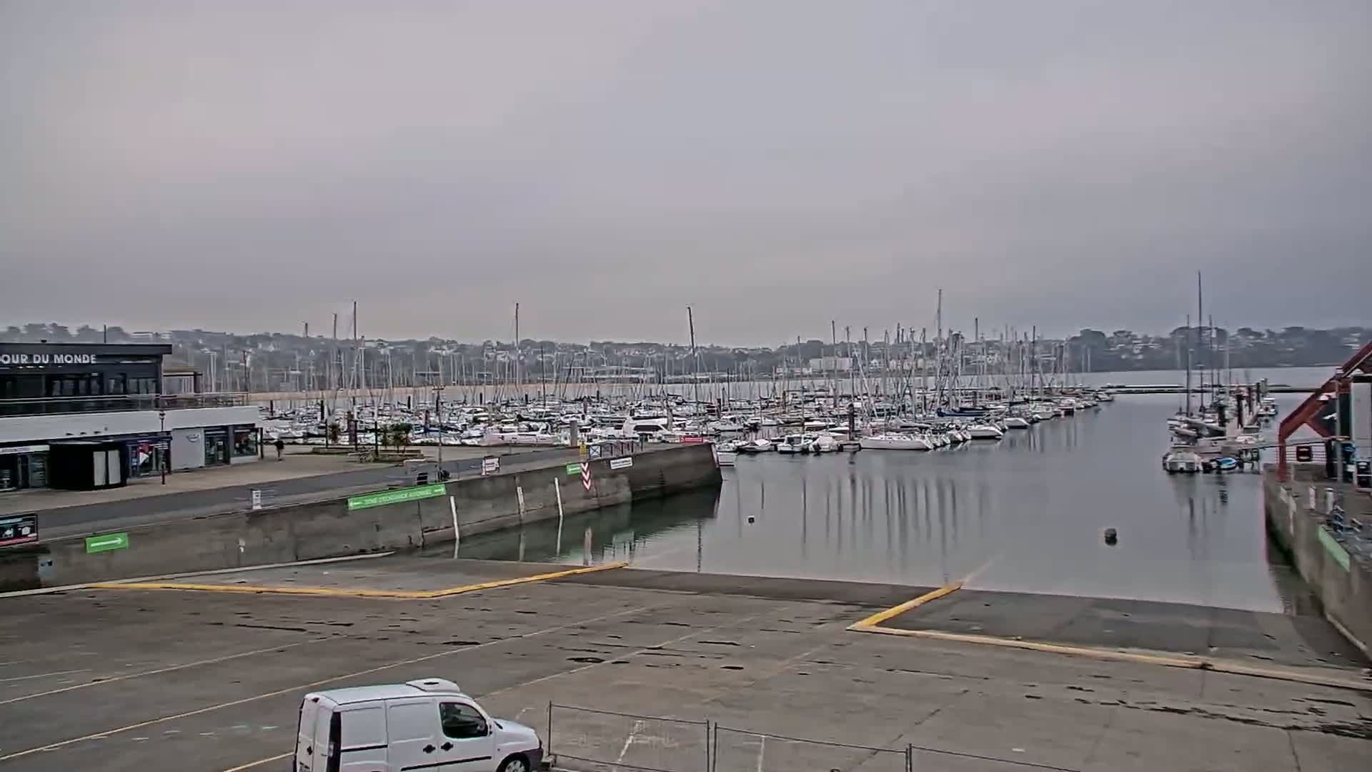 A marina filled with numerous sailboats under an overcast sky, with a van parked in an empty parking lot nearby.