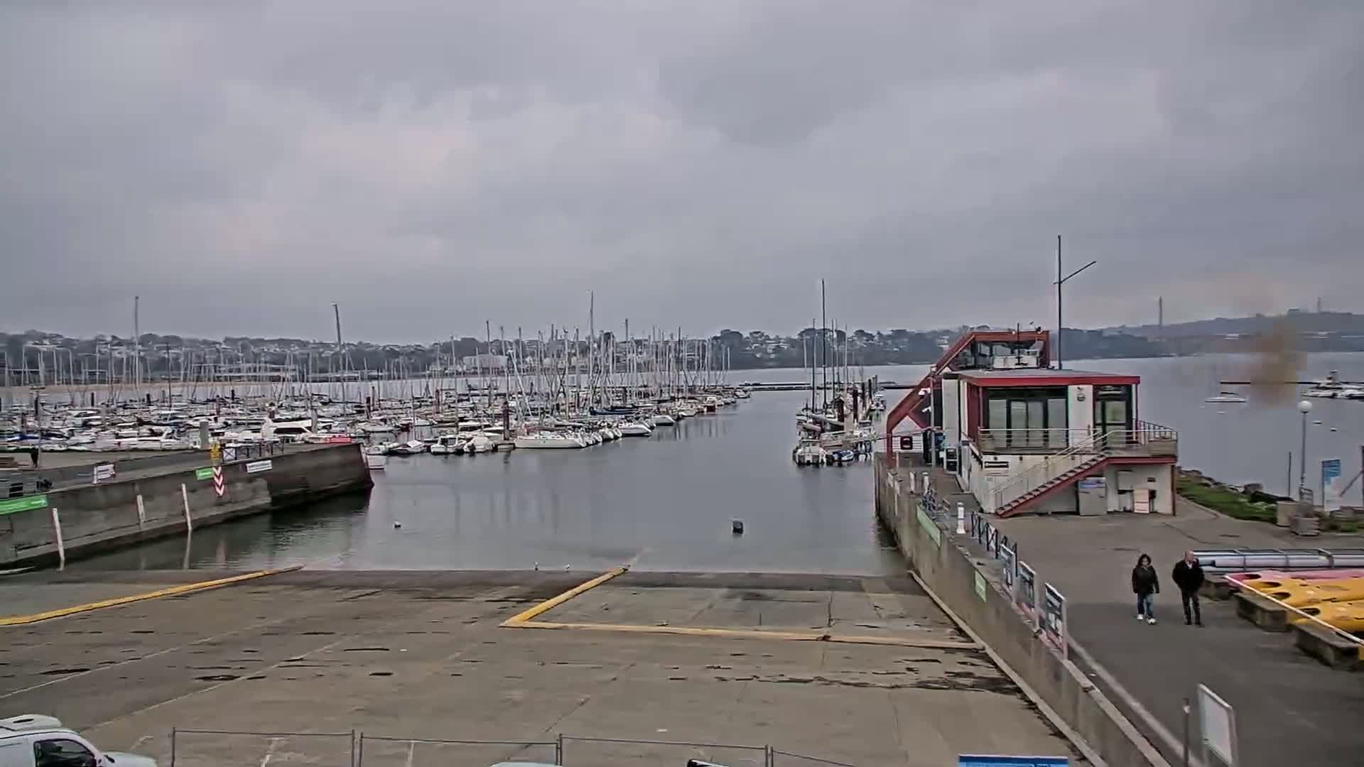 A marina filled with numerous sailboats is seen on an overcast day, with a small building and a boat ramp in the foreground.