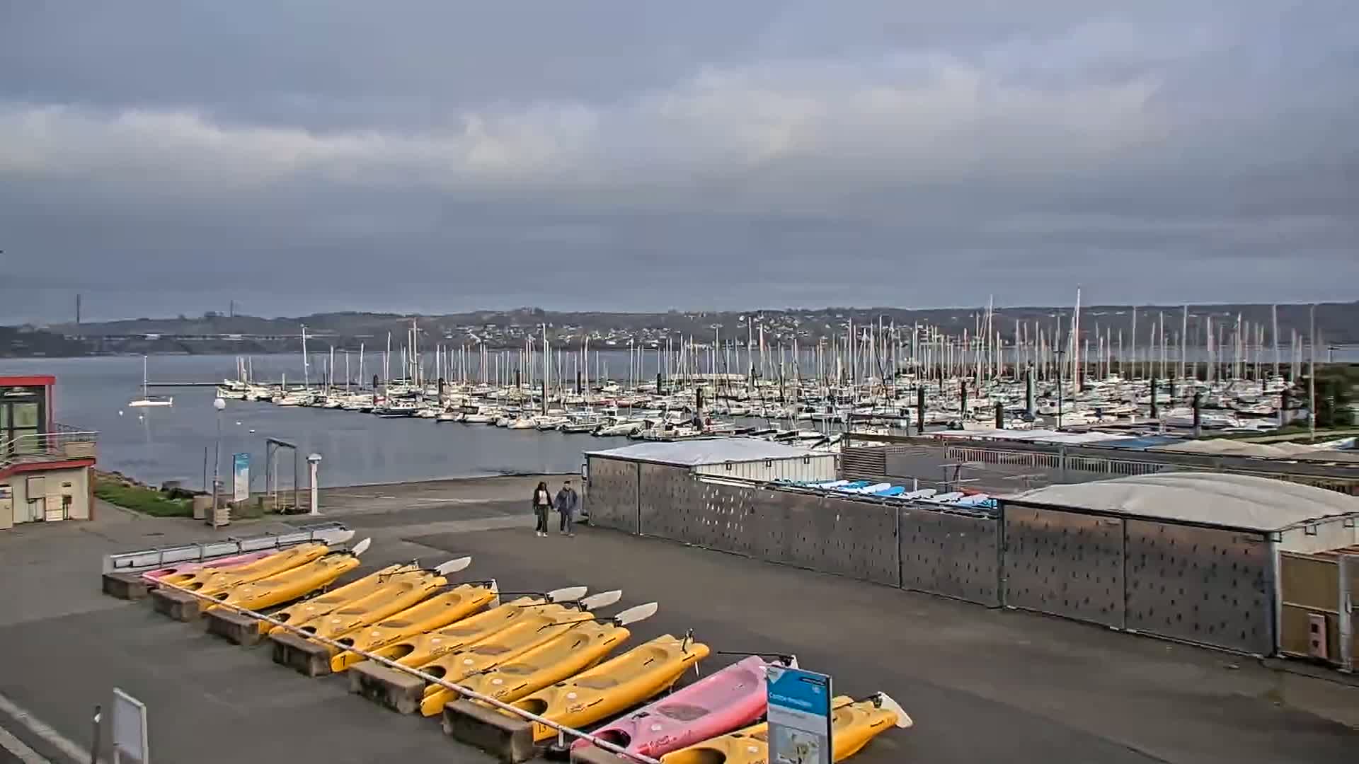 A marina filled with sailboats under a cloudy sky, with kayaks lined up on the shore next to a couple walking by.