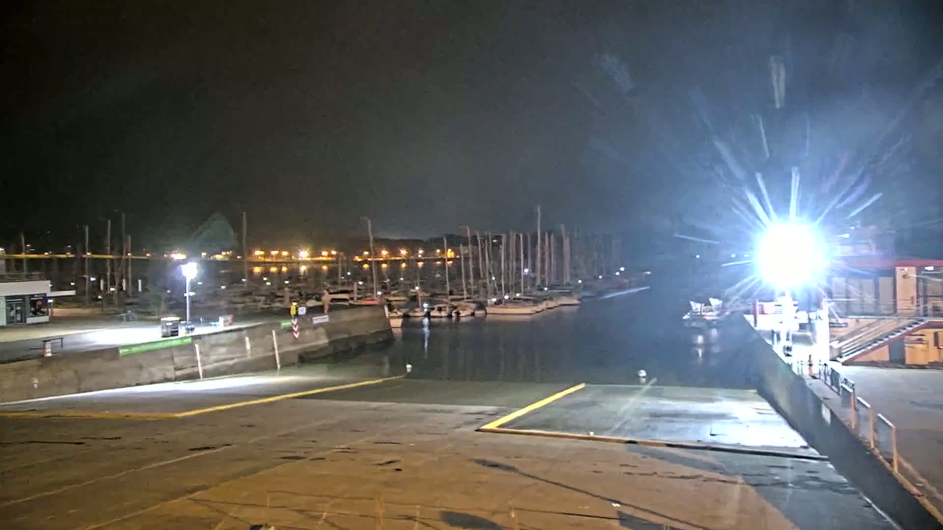 A brightly lit boat launch at night overlooks a calm harbor filled with docked sailboats under a dark sky.