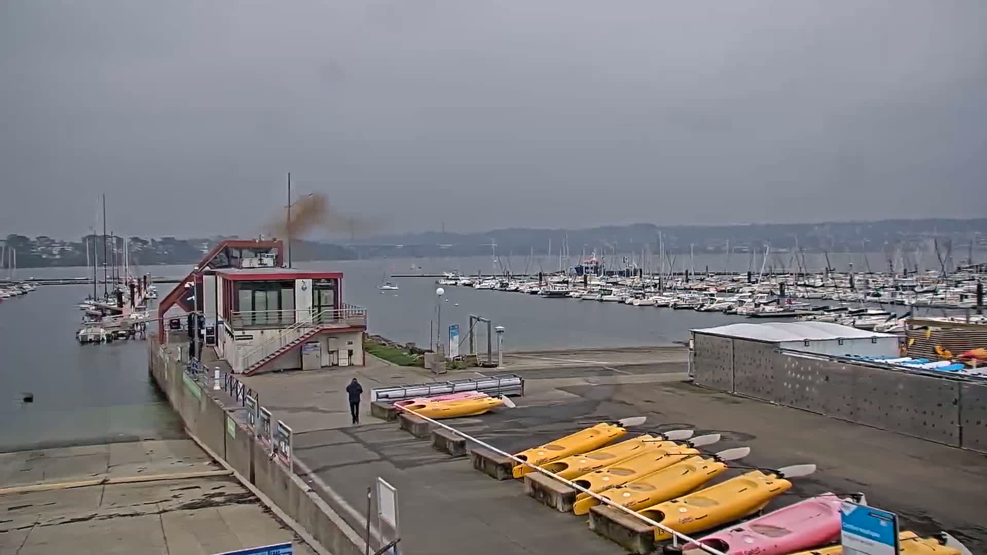 A marina with numerous boats, a small building, and a row of kayaks is visible under an overcast sky.