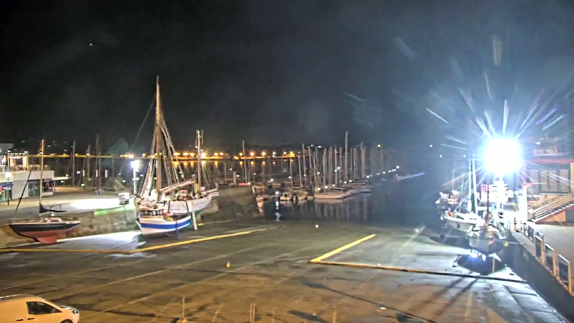 At night, several sailboats are docked in a harbor under bright artificial lights, with additional boats visible in the background.