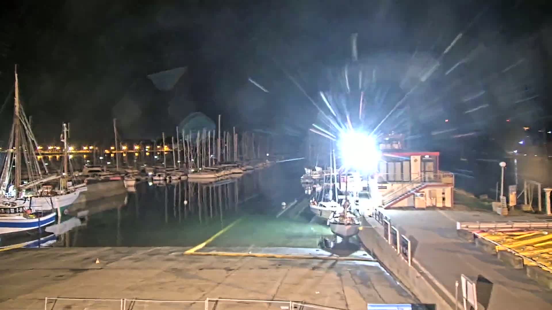 At night, a brightly lit marina shows numerous boats docked, with several more boats near a dockside building under intense artificial light in what appears to be windy conditions.