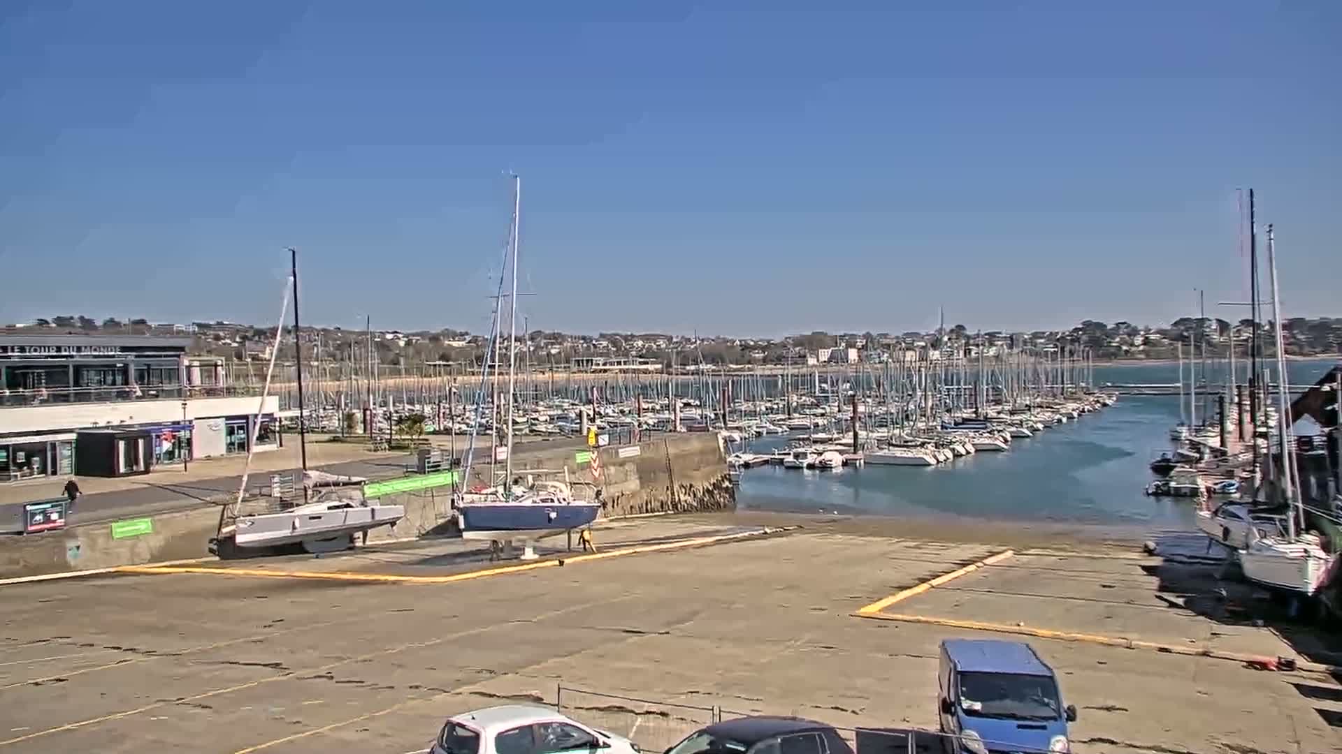 A marina filled with numerous sailboats under a clear blue sky, with a building and parking area in the foreground.