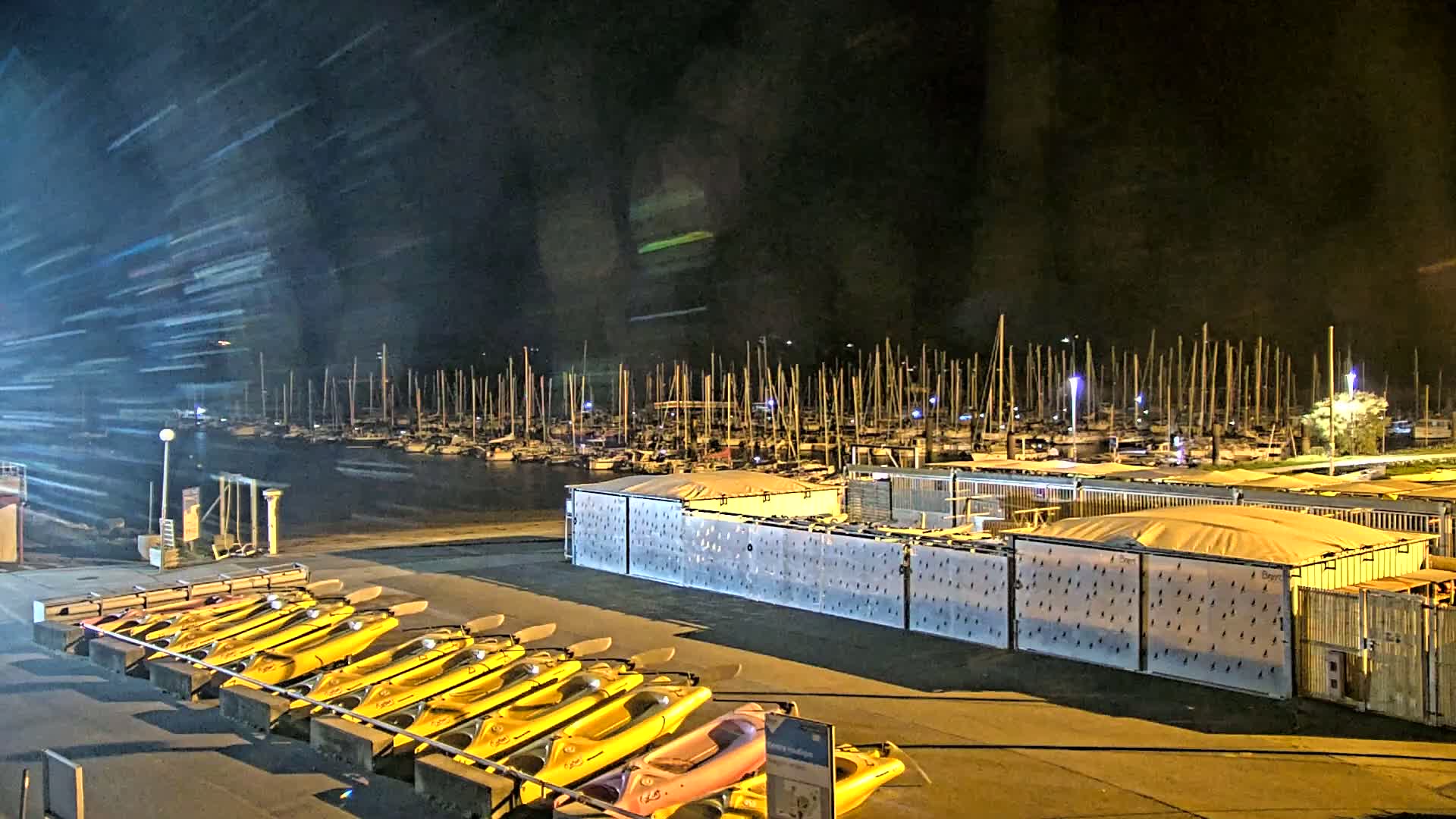 At night, under a dark sky with streaks of light, numerous yellow kayaks are lined up near a marina full of boats, behind a fenced-in storage area.