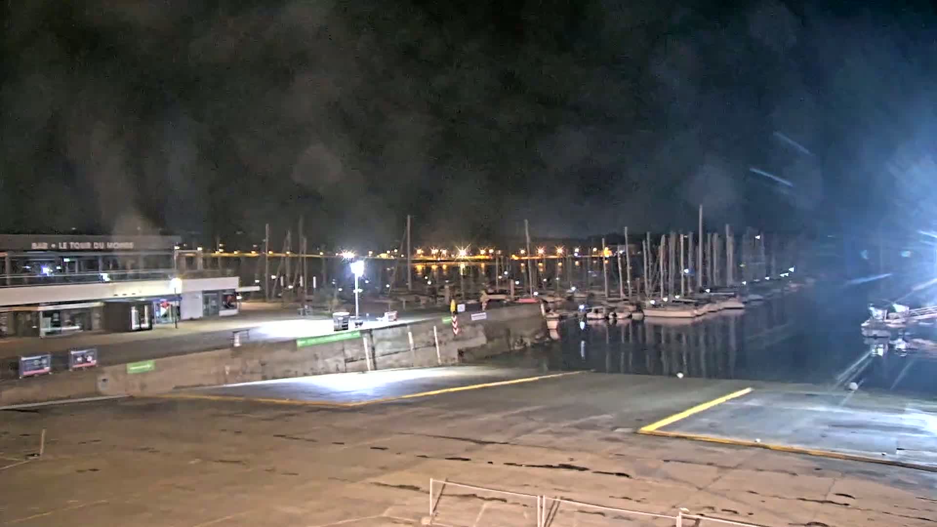 A nighttime view of a marina with numerous boats docked, under a dark, cloudy sky.